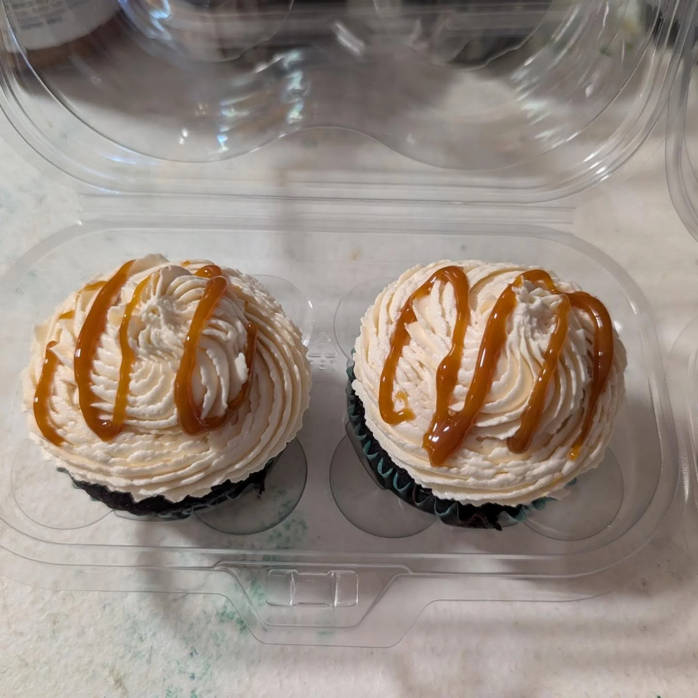 Two chocolate cupcakes with creamy frosting drizzled with caramel sauce, presented in a plastic container.