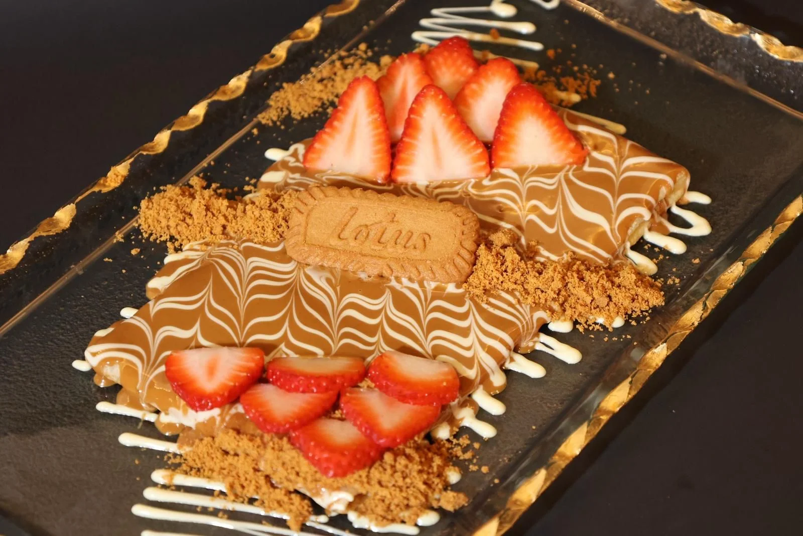 A dessert platter featuring sweet toppings with chocolate, white drizzles, strawberries, and cookie crumbs. The dish is elegantly arranged.