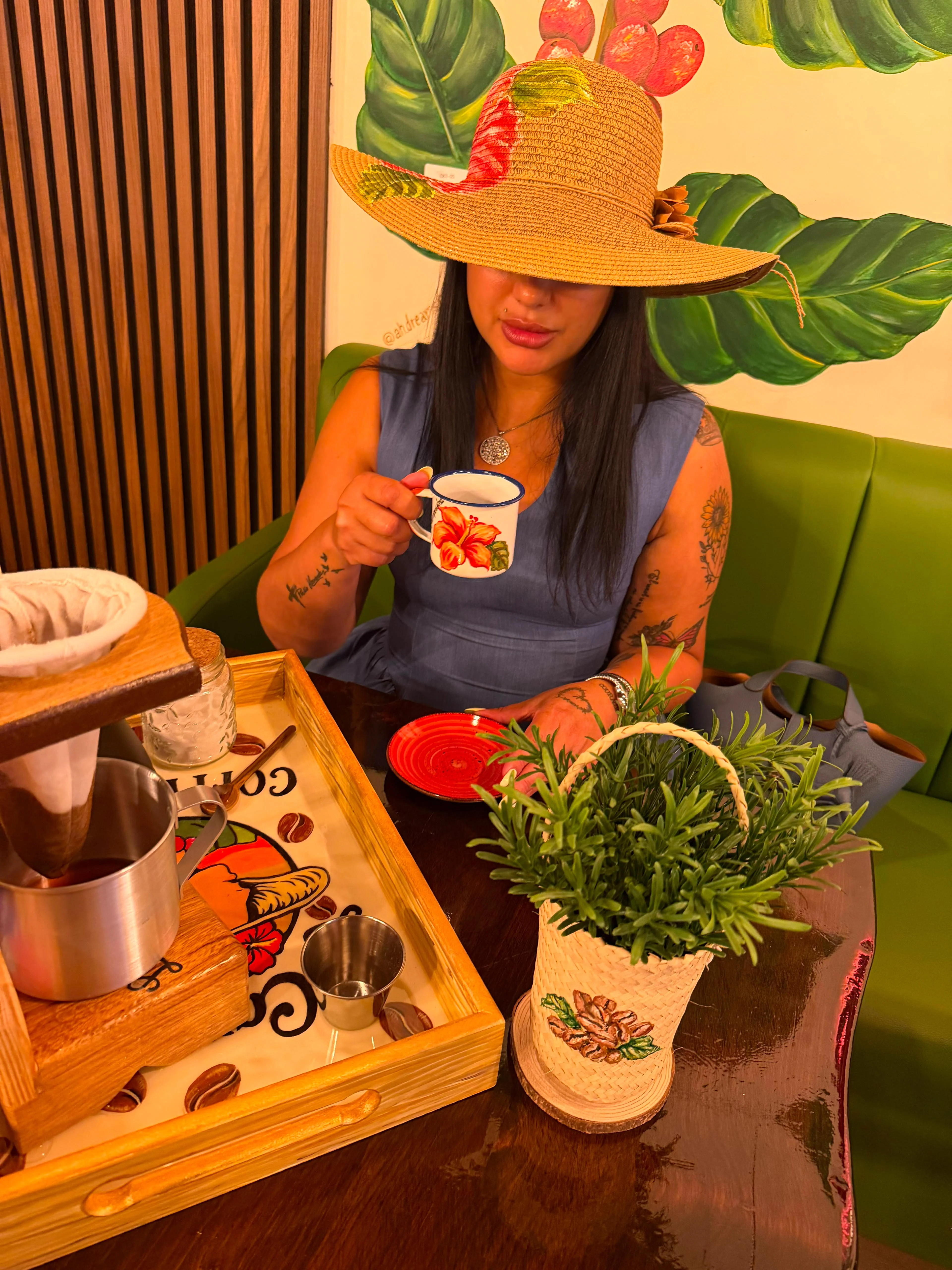 A woman in a wide-brimmed hat holds a cup in a vividly decorated cafe with plants and coffee-making tools.