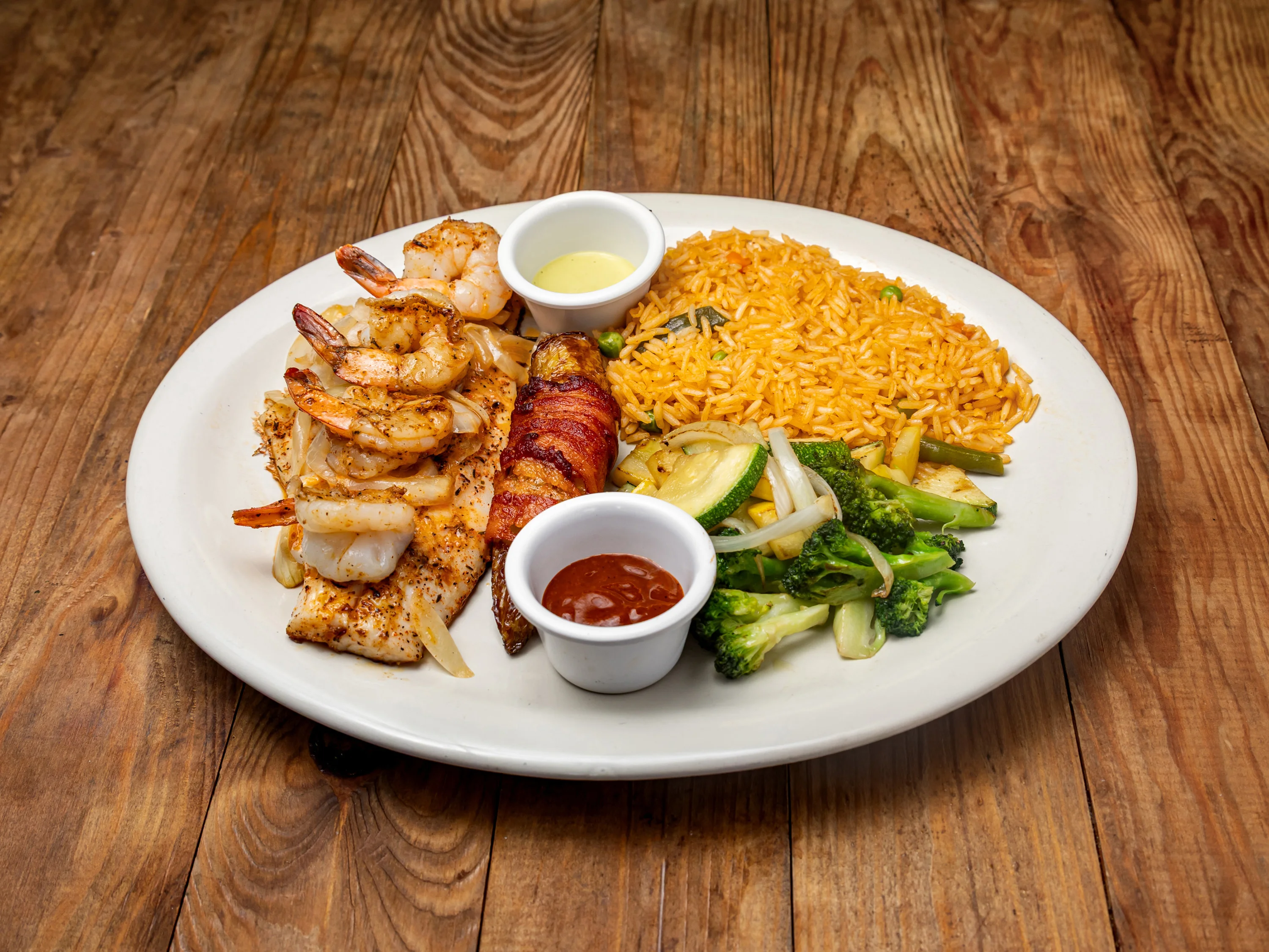 A seafood platter featuring grilled shrimp, fish, and vegetables, served alongside yellow rice and various dipping sauces.