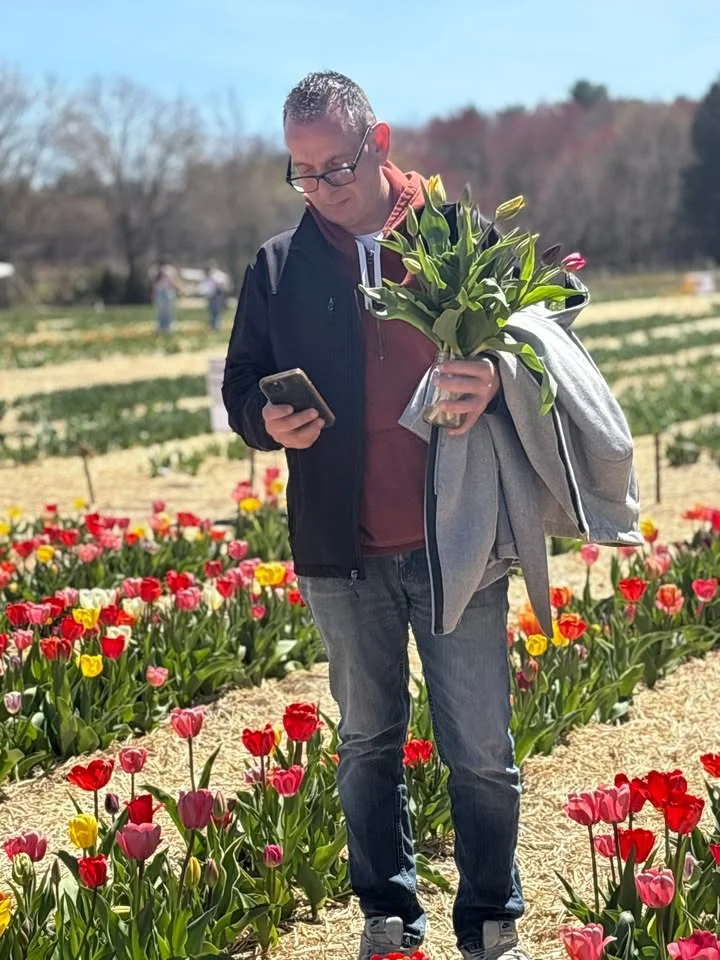 A person standing in a colorful flower field, holding a bouquet of tulips and a phone while wearing a jacket.