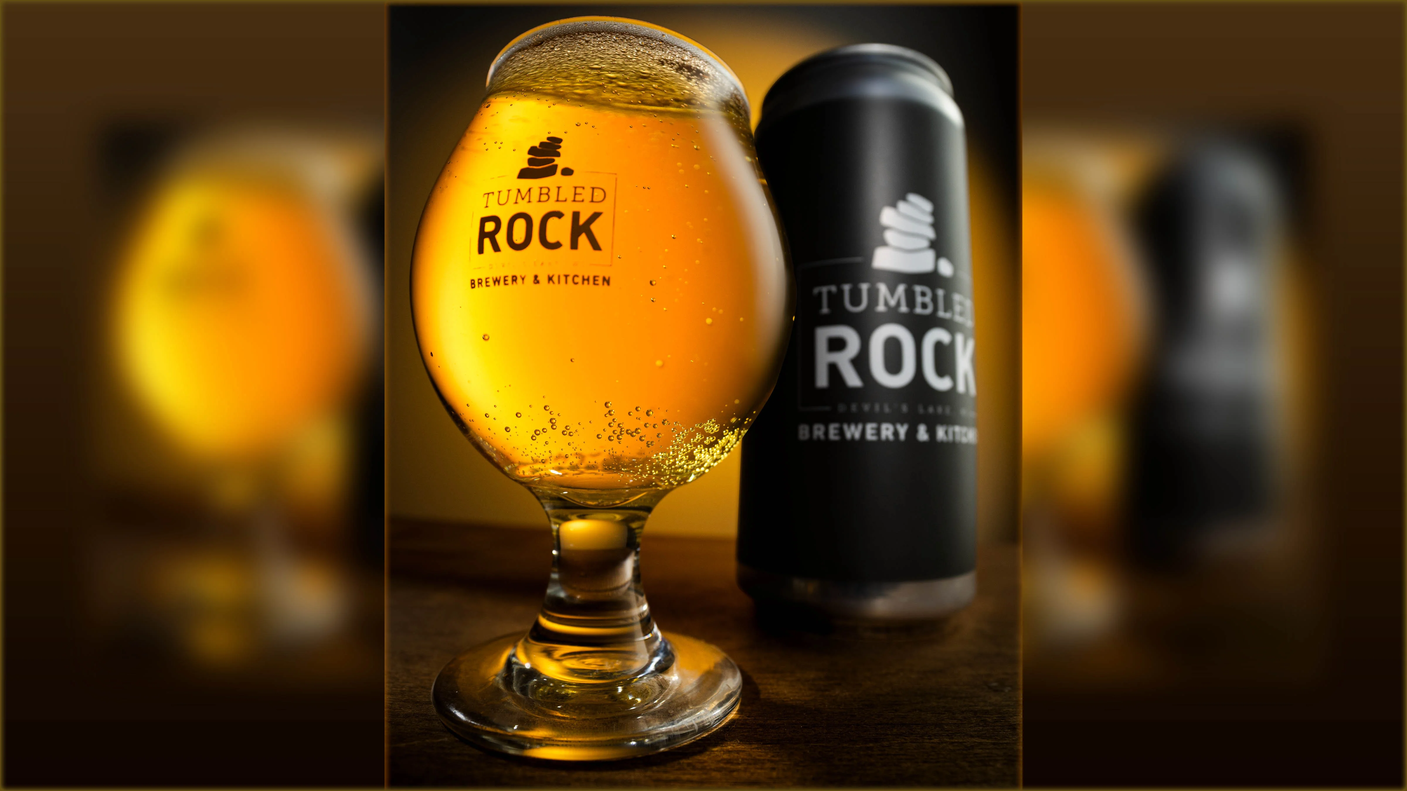 A glass of beer with bubbles next to a can, both branded Tumbled Rock Brewery.