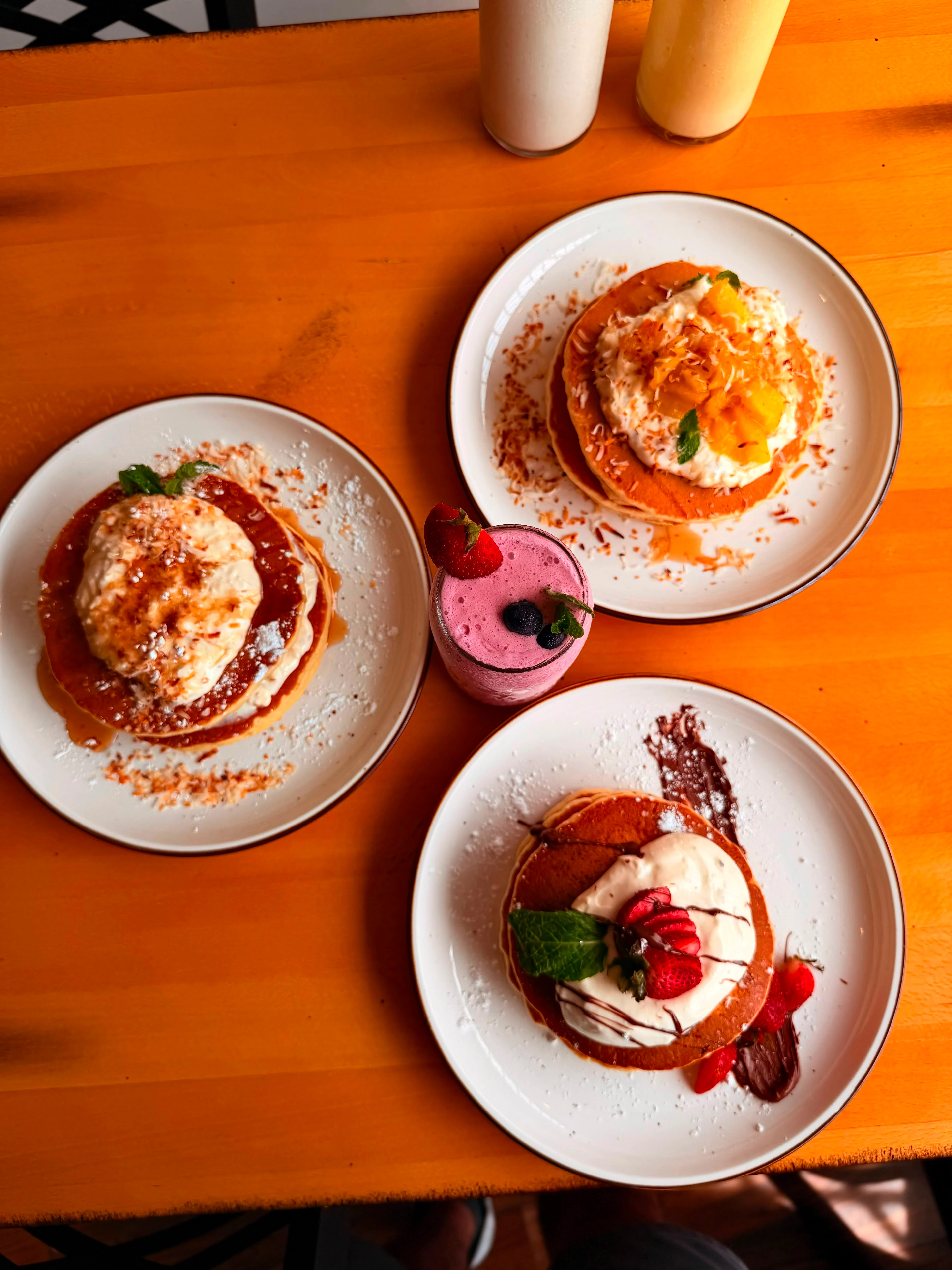 A plate of three fluffy pancakes topped with whipped cream, strawberries, and a purple edible flower, accompanied by a small bowl of mixed fruits.
