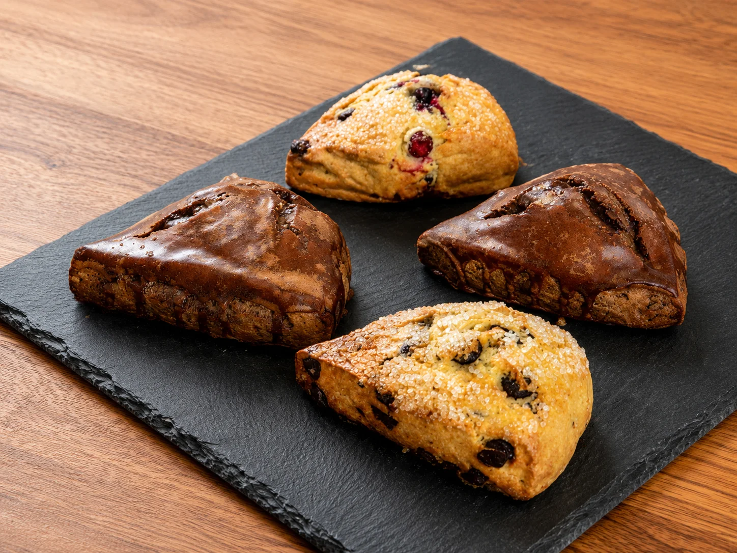 Four assorted scones on a slate board, featuring chocolate and fruit flavors.