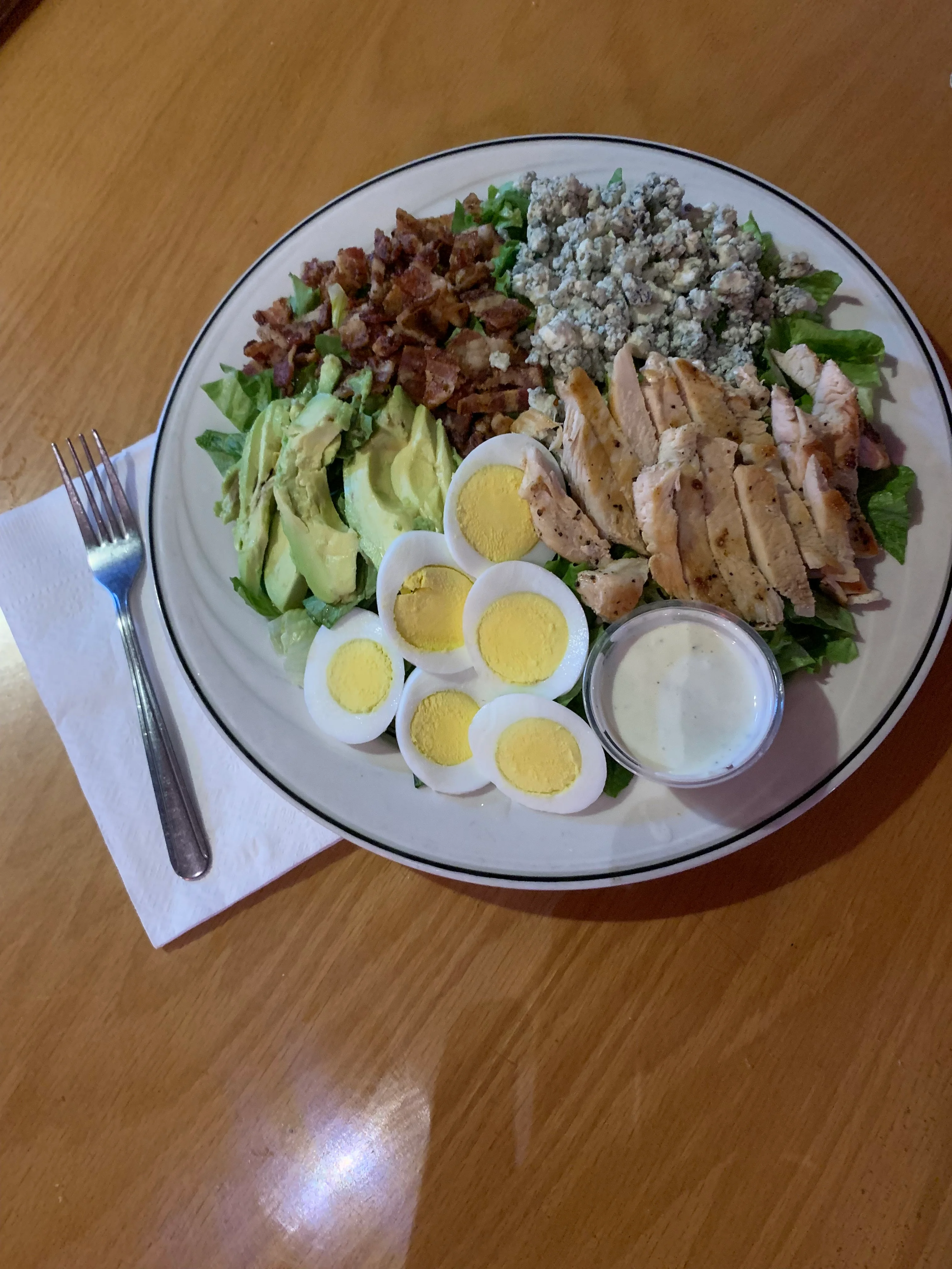 A fresh salad topped with grilled chicken, bacon, avocado, blue cheese, and hard-boiled eggs, served with a side of ranch dressing.