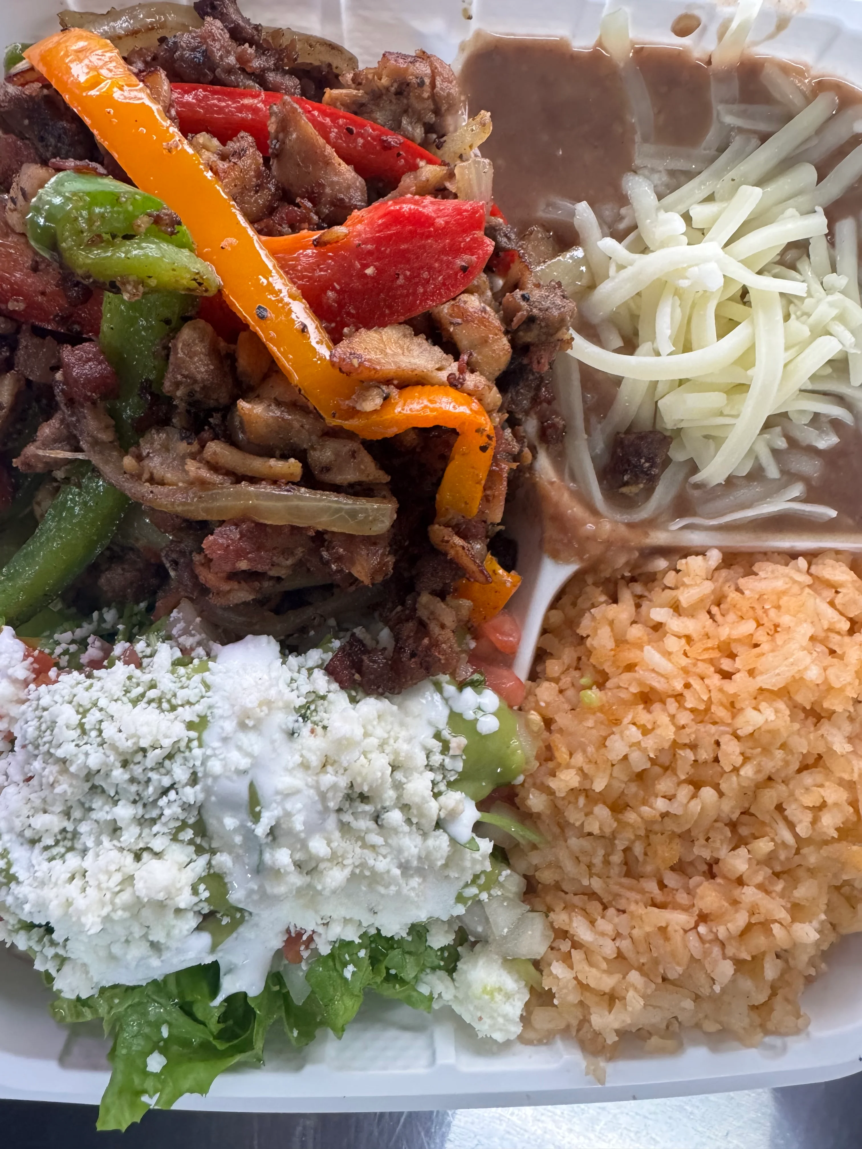 A plate with grilled meat and peppers served alongside rice, refried beans, and fresh vegetables.