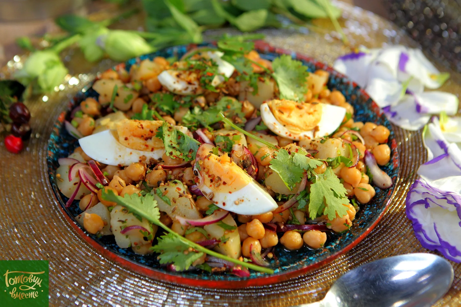 A colorful salad featuring boiled eggs, chickpeas, diced potatoes, and fresh herbs, elegantly arranged on a decorative plate, surrounded by petals and greens.