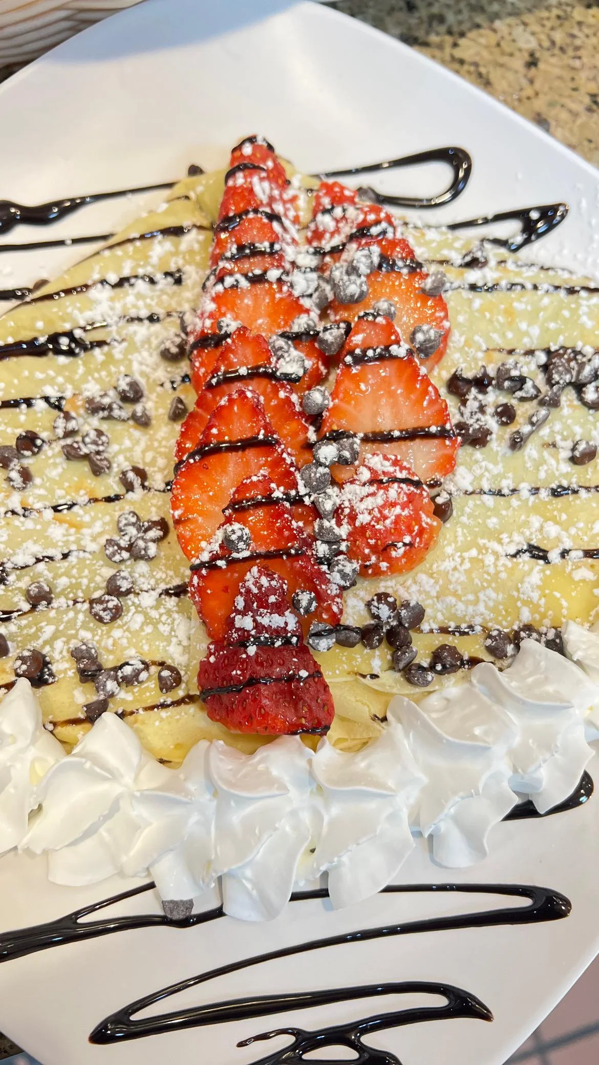 A delicious crepe topped with fresh strawberries, chocolate drizzle, powdered sugar, and whipped cream, presented on a square plate.
