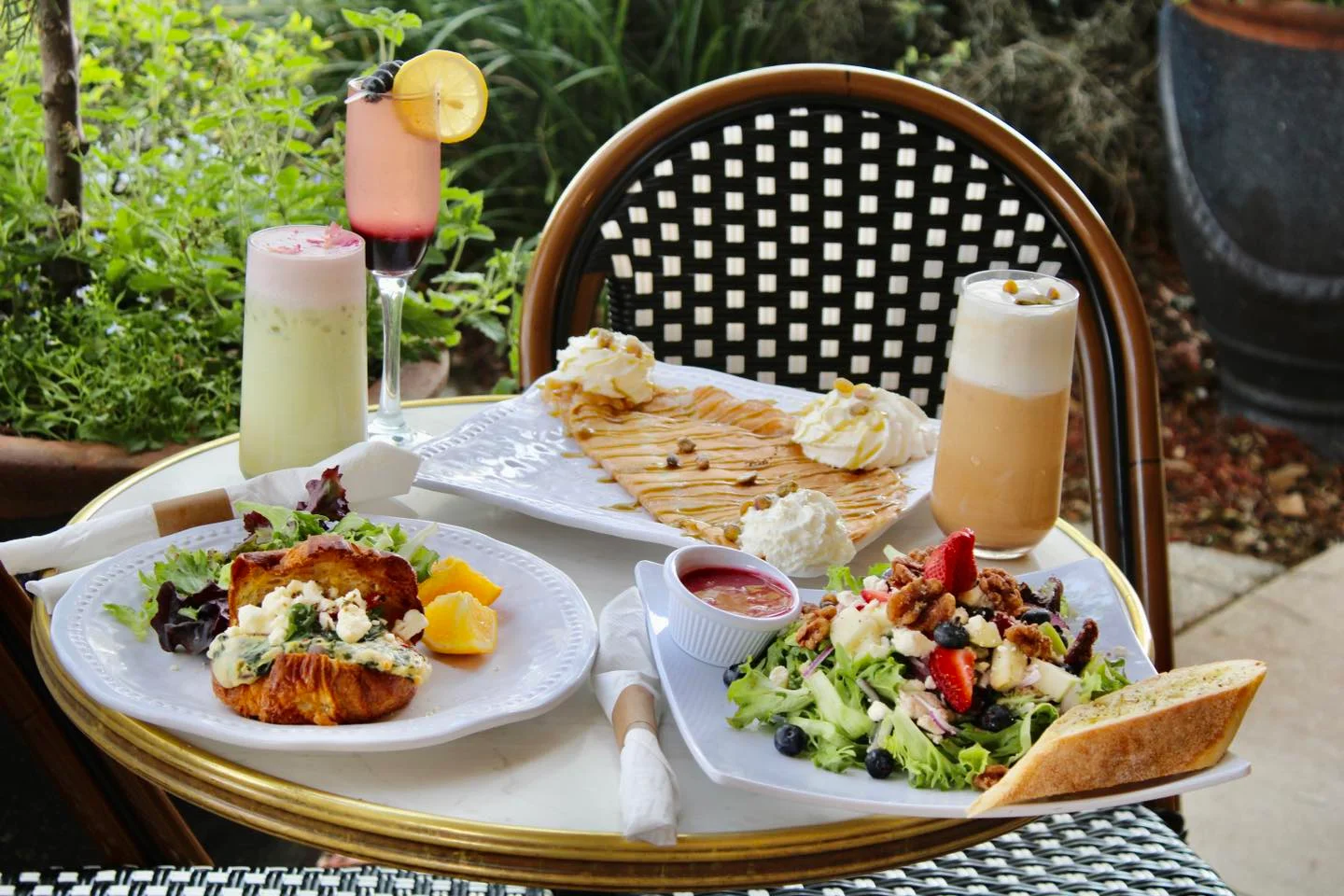A cozy table setting featuring a plate with a savory croissant dish, a fresh salad, and delicious drinks, surrounded by greenery for a relaxed atmosphere.