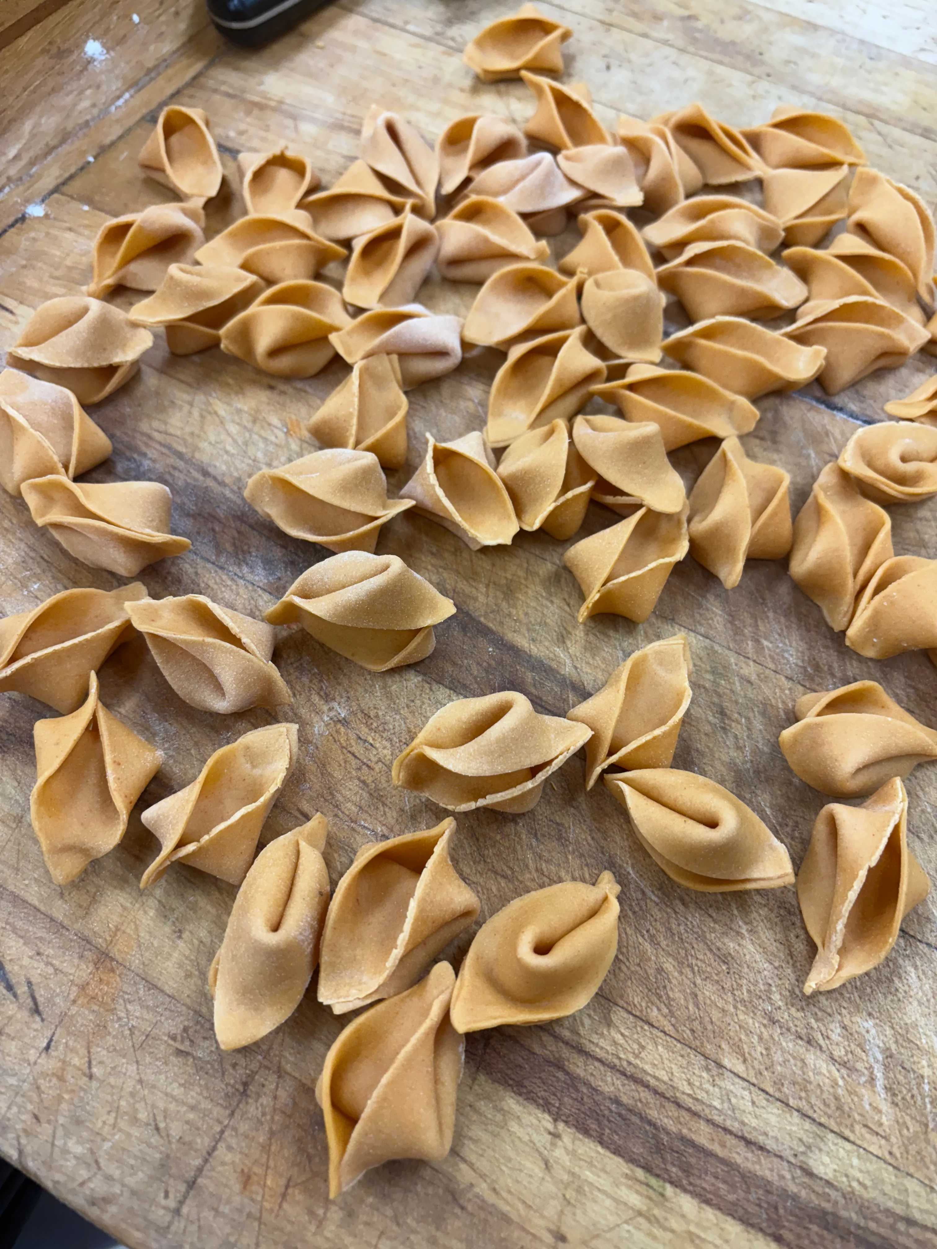 A collection of uncooked pasta pieces scattered on a wooden surface. The pasta is shaped like small shells with a smooth texture.