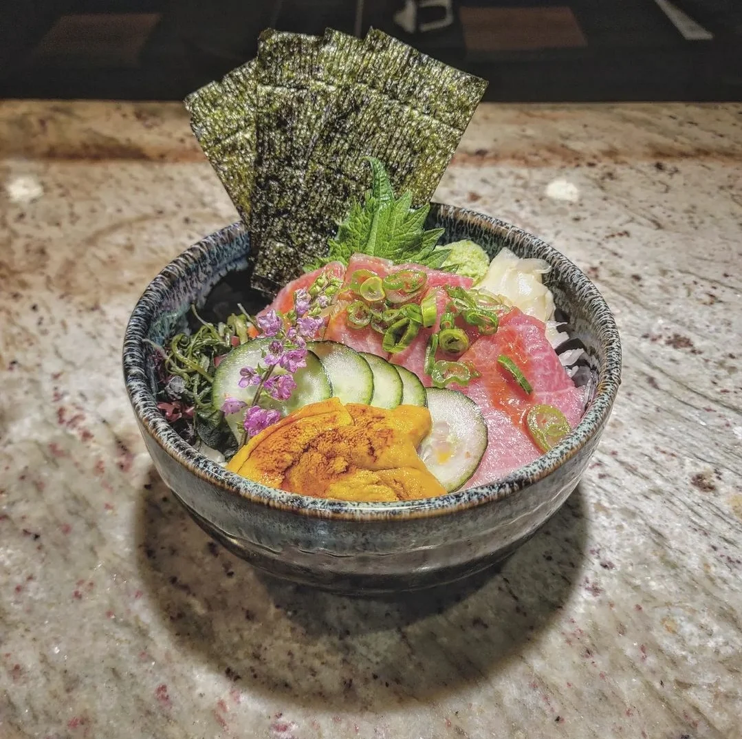 A beautifully presented sushi dish featuring rice, various toppings including fried elements, sliced lotus root, and garnishes drizzled with a creamy sauce.