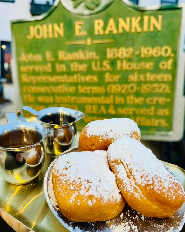 A tray featuring three powdered donuts alongside two small cups of dipping sauce. The background includes a historical plaque.