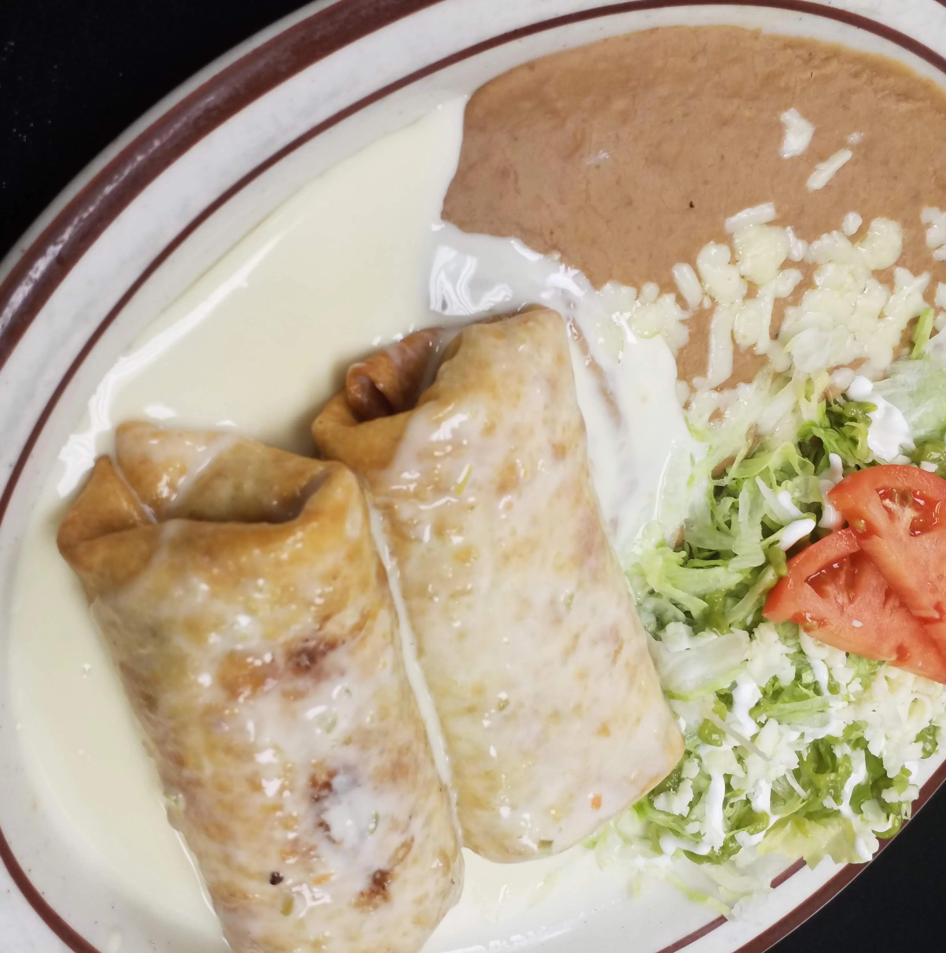 The best golden-fried chimichangas in Radford topped with creamy cheese sauce, served with a side of refried beans garnished with melted cheese and a fresh salad of shredded lettuce, sour cream, and tomato slices.