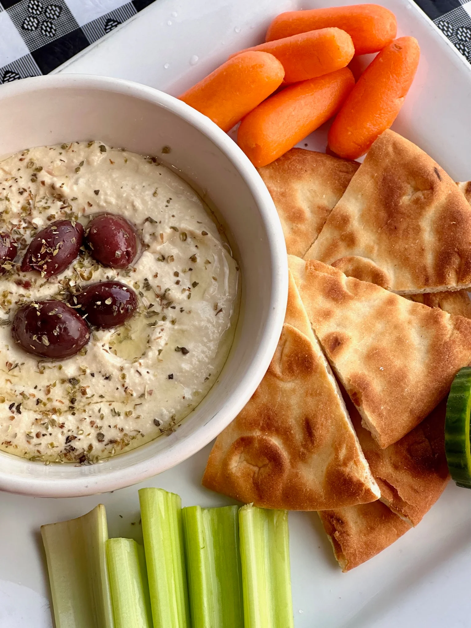 A bowl of hummus topped with olives, accompanied by pita bread, carrots, celery, and cucumber slices.