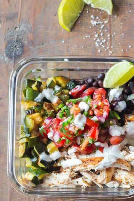 A healthy meal prep container filled with grilled chicken, black beans, fresh salsa, and sautéed vegetables, garnished with lime wedges.