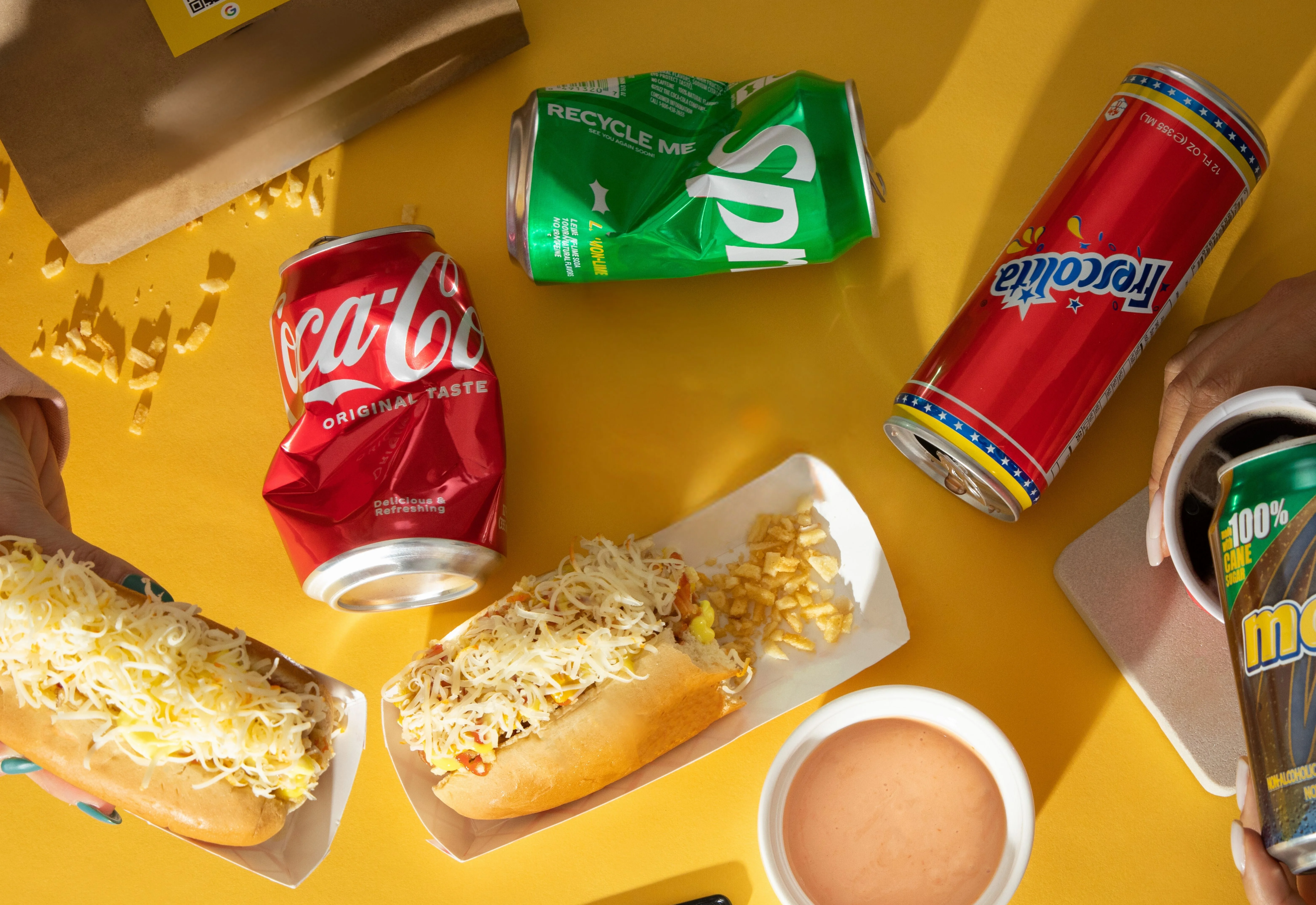 Two loaded hot dogs with drinks and dipping sauce on a yellow background.