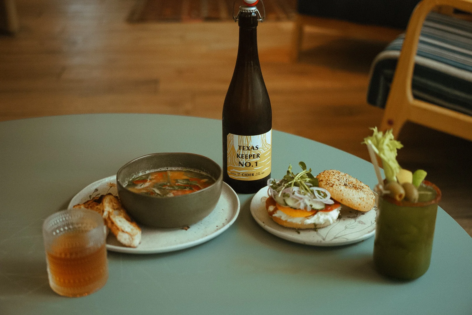 A table setting featuring a bowl of soup, a bottle of cider, a bagel with toppings, and a cocktail glass. The scene is cozy with wooden flooring and casual decor.