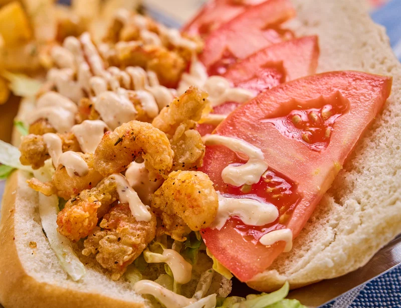 A sandwich featuring crispy fried shrimp, fresh tomato slices, and a drizzle of creamy sauce on a bed of lettuce, served on soft white bread.