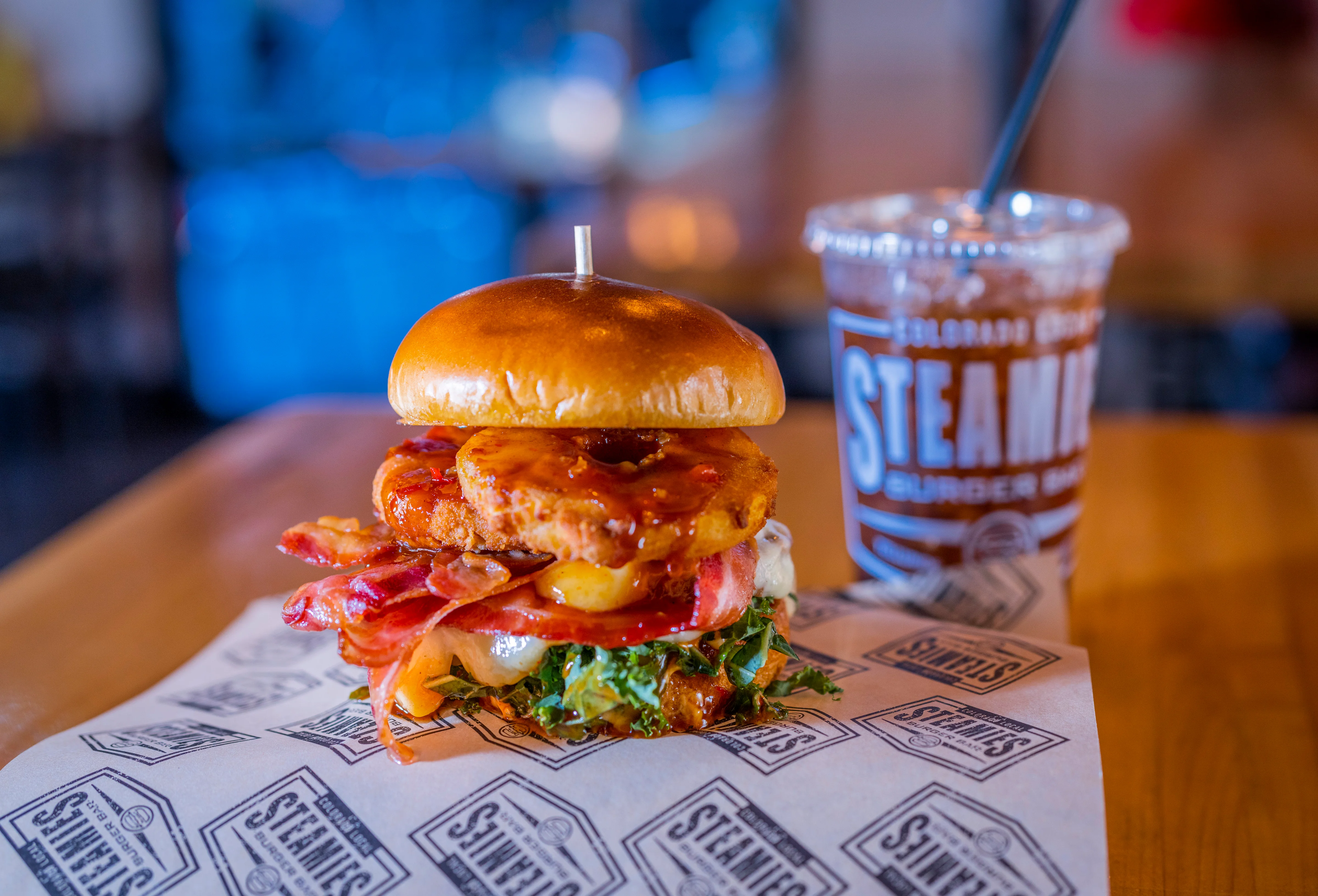 A stacked burger featuring bacon, crispy chicken, and pineapple with a toasted bun. It sits on printed parchment paper next to a drink.