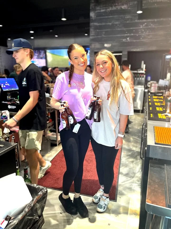 Two bartenders smiling with drinks at a lively bar