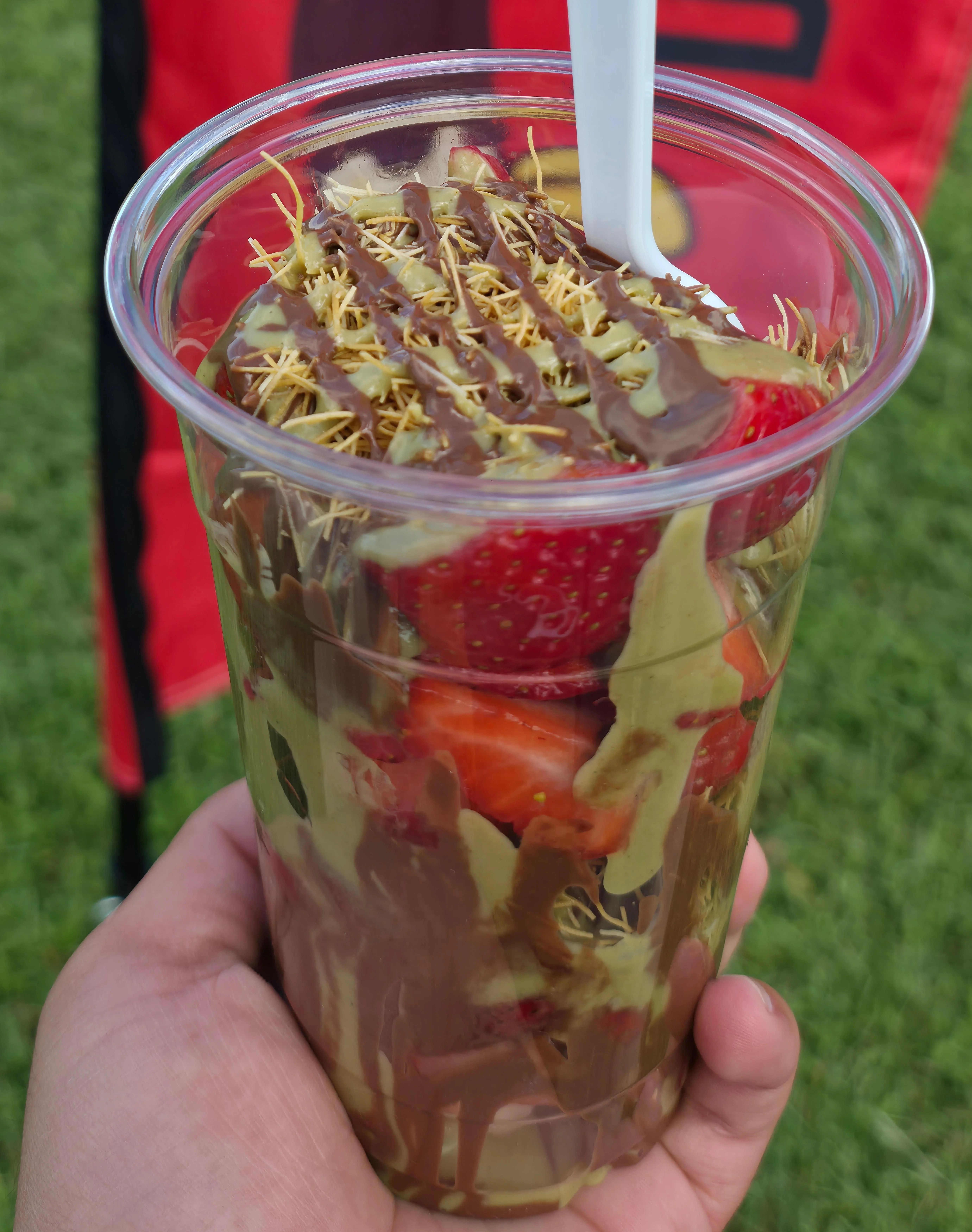 A hand holding a plastic cup filled with strawberries and a swirl of chocolate, topped with a decorative layer, set against a grassy backdrop.