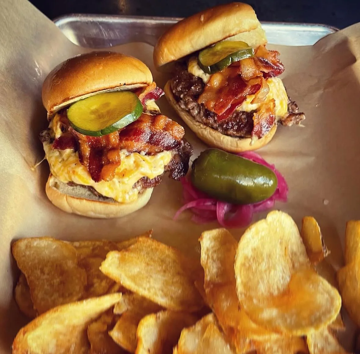 Two grilled burgers topped with bacon and pickles, served alongside a side of crispy potato chips.