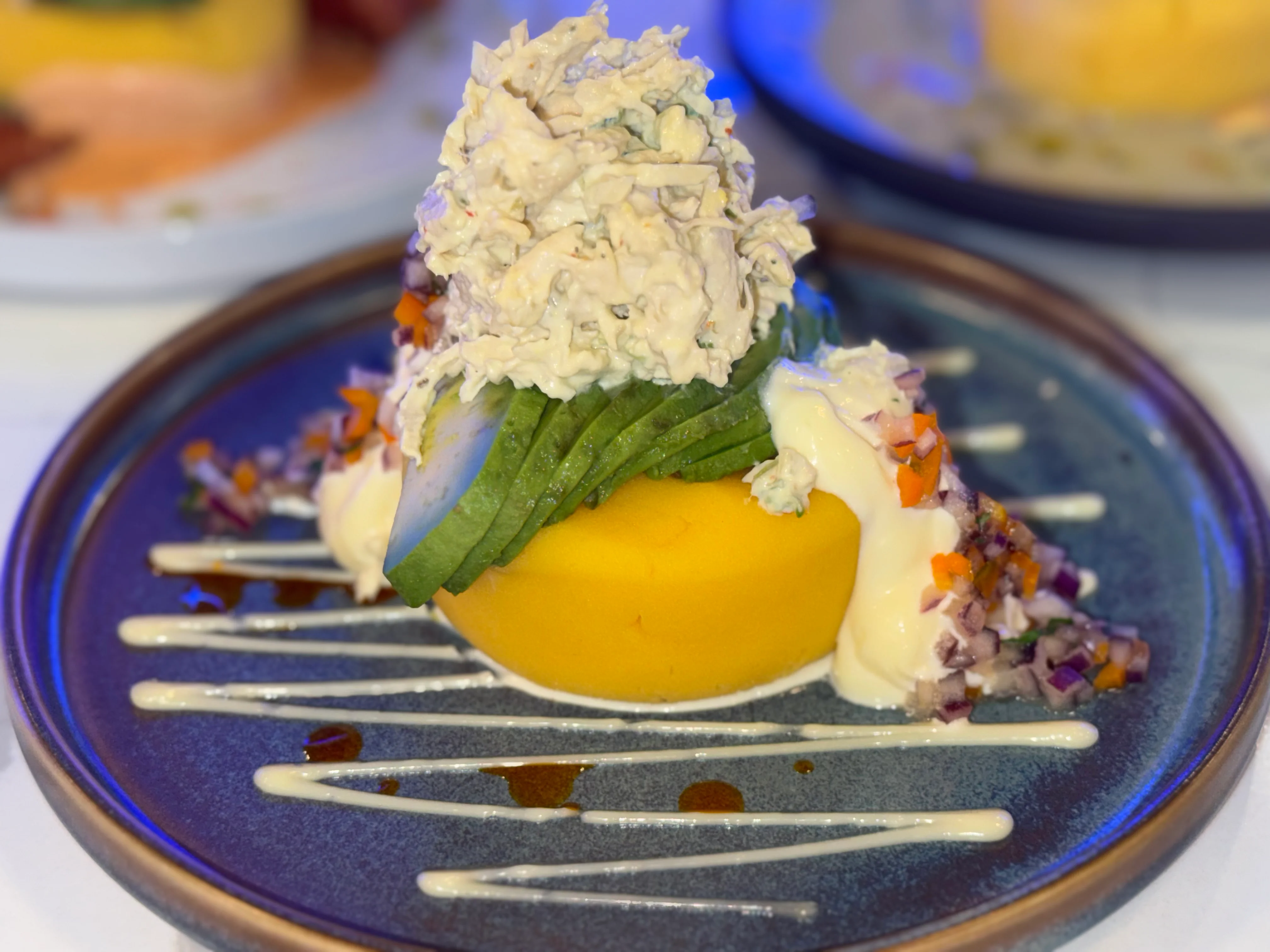 A vibrant dish featuring a mound of shredded chicken on top of a yellow circular base, garnished with avocado slices, diced vegetables, and drizzled with a creamy sauce.