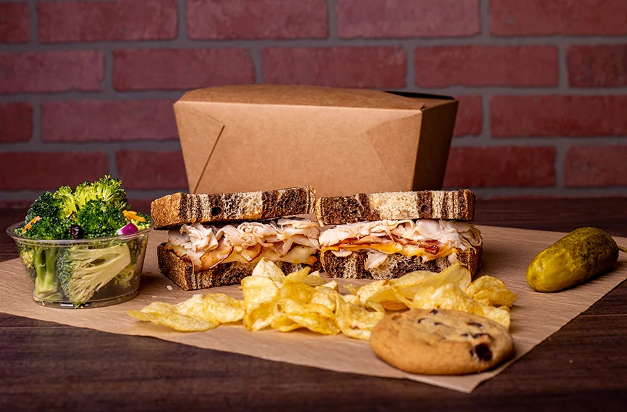 A boxed meal featuring a thick turkey and bacon sandwich on marbled rye bread, served with a side of potato chips, a chocolate chip cookie, a small cup of fresh broccoli salad, and a whole pickle. A brown takeout box sits in the background against a brick wall backdrop.