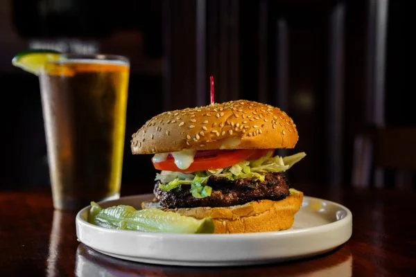 A burger served with pickles and greens, garnished with a knife and a side of sauce.