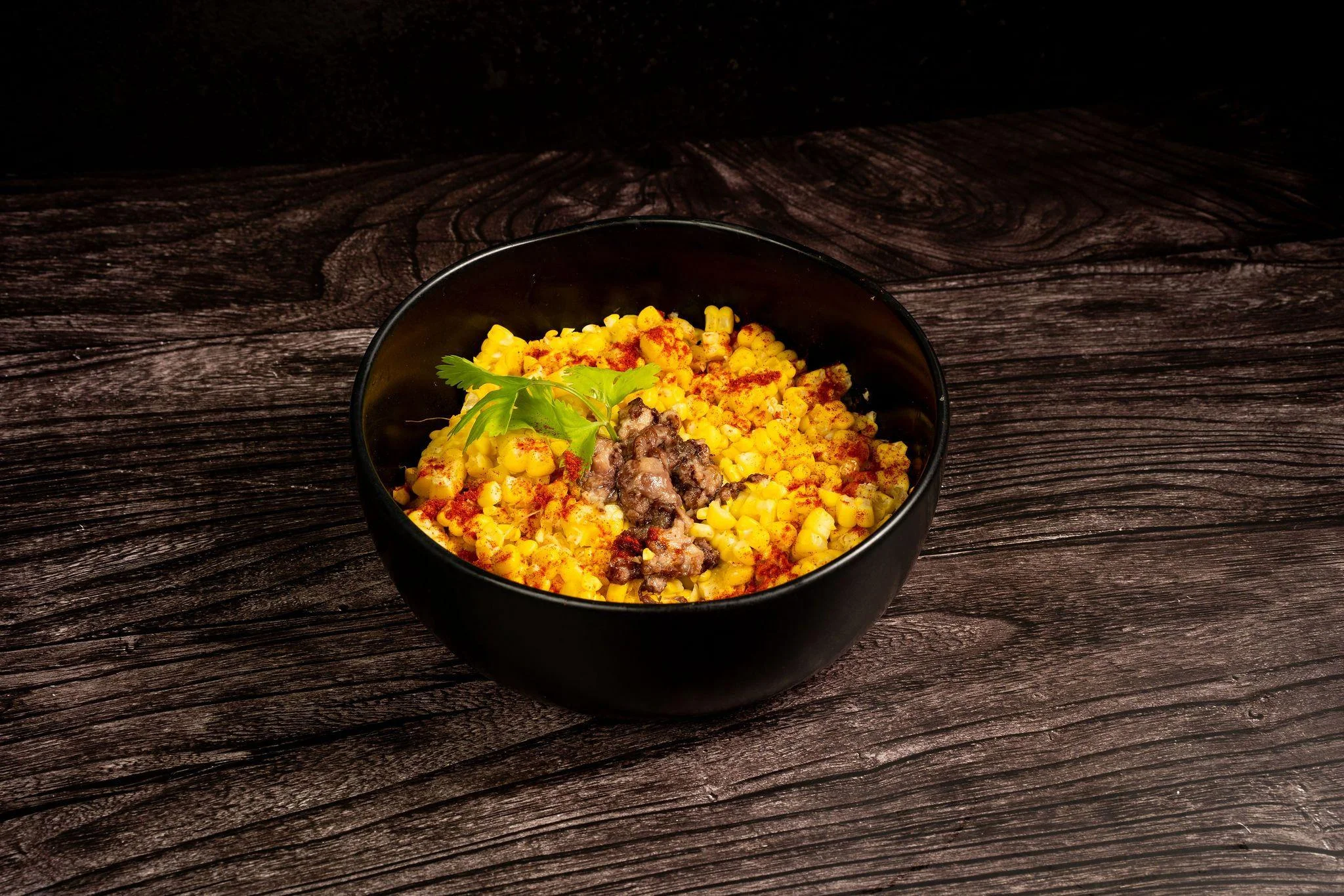 A black bowl filled with yellow corn and seasoned meat, garnished with herbs.