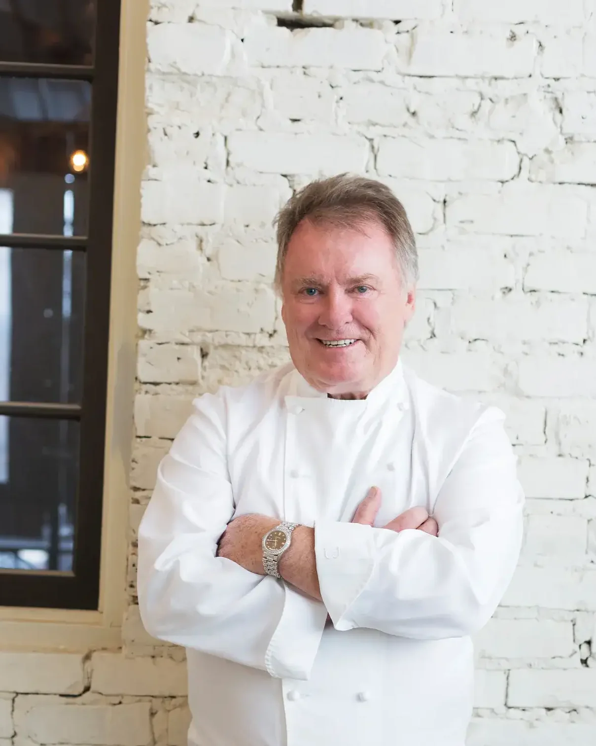 Chef in a white coat smiling against a brick wall