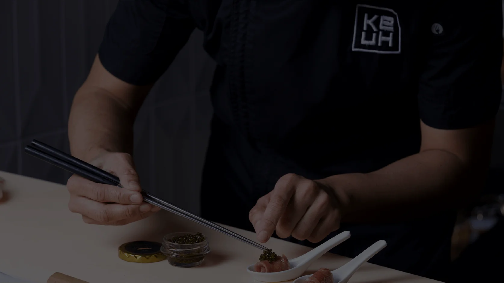 A chef wearing a branded Ke-uH uniform uses a brush to apply sauce from a stainless steel container onto small appetizers served in individual white ceramic spoons.