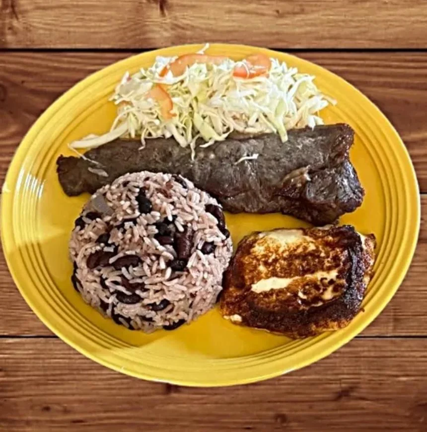 A colorful plate featuring a portion of grilled meat, rice with black beans, a side of cabbage salad, and a slice of fried cheese, all arranged on a yellow plate.