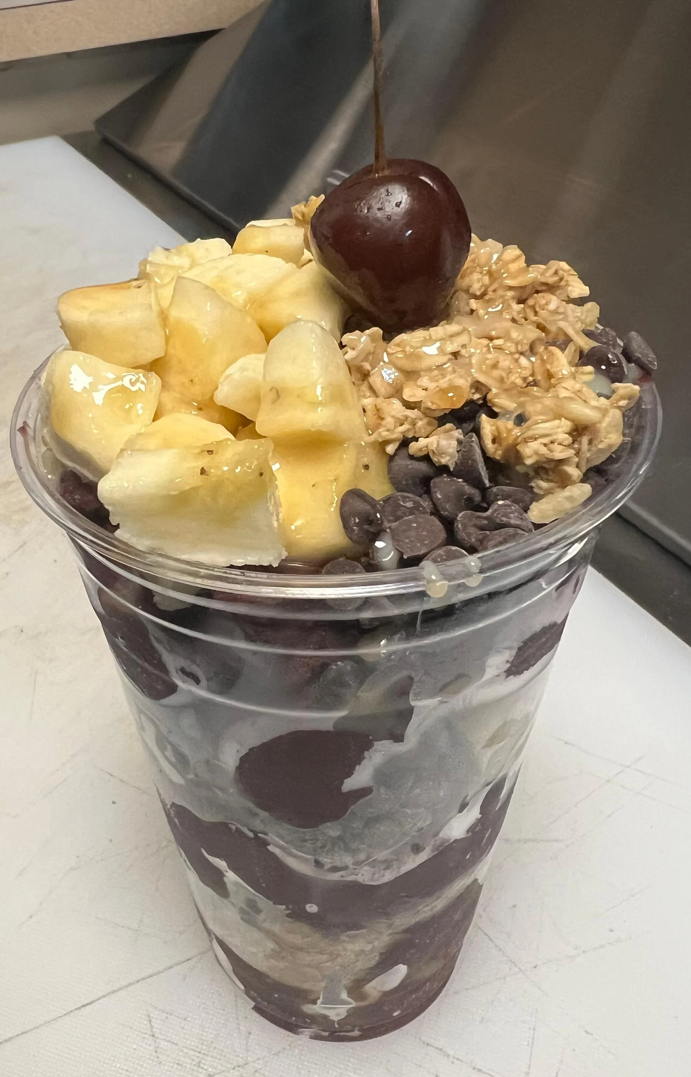 A layered acai bowl topped with banana chunks, granola, chocolate chips, and a cherry on top, presented in a clear cup.