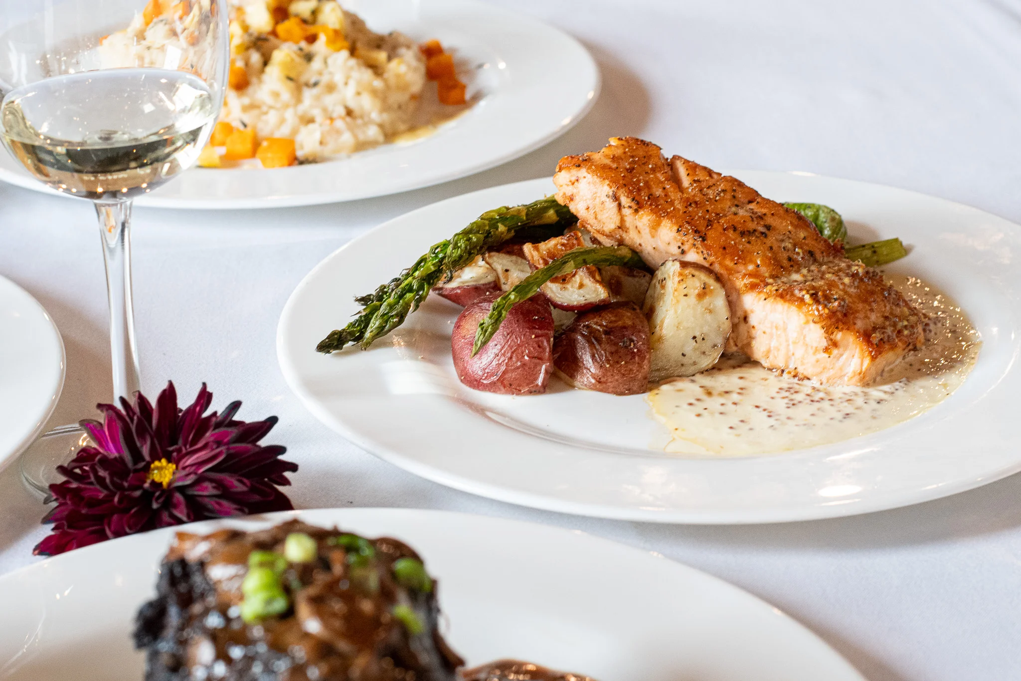 A gourmet dish featuring grilled salmon served with roasted potatoes and asparagus, alongside a glass of white wine and a decorative flower.