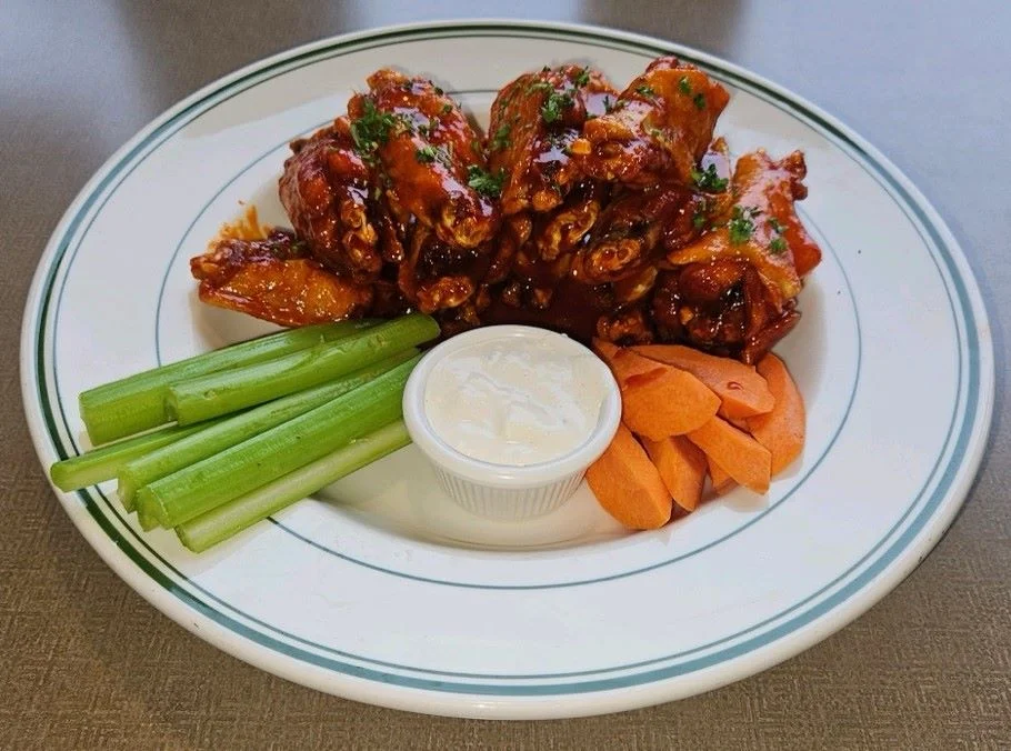 A plate of chicken wings coated in a sticky sauce, accompanied by celery, carrot sticks, and a small dish of ranch dressing.