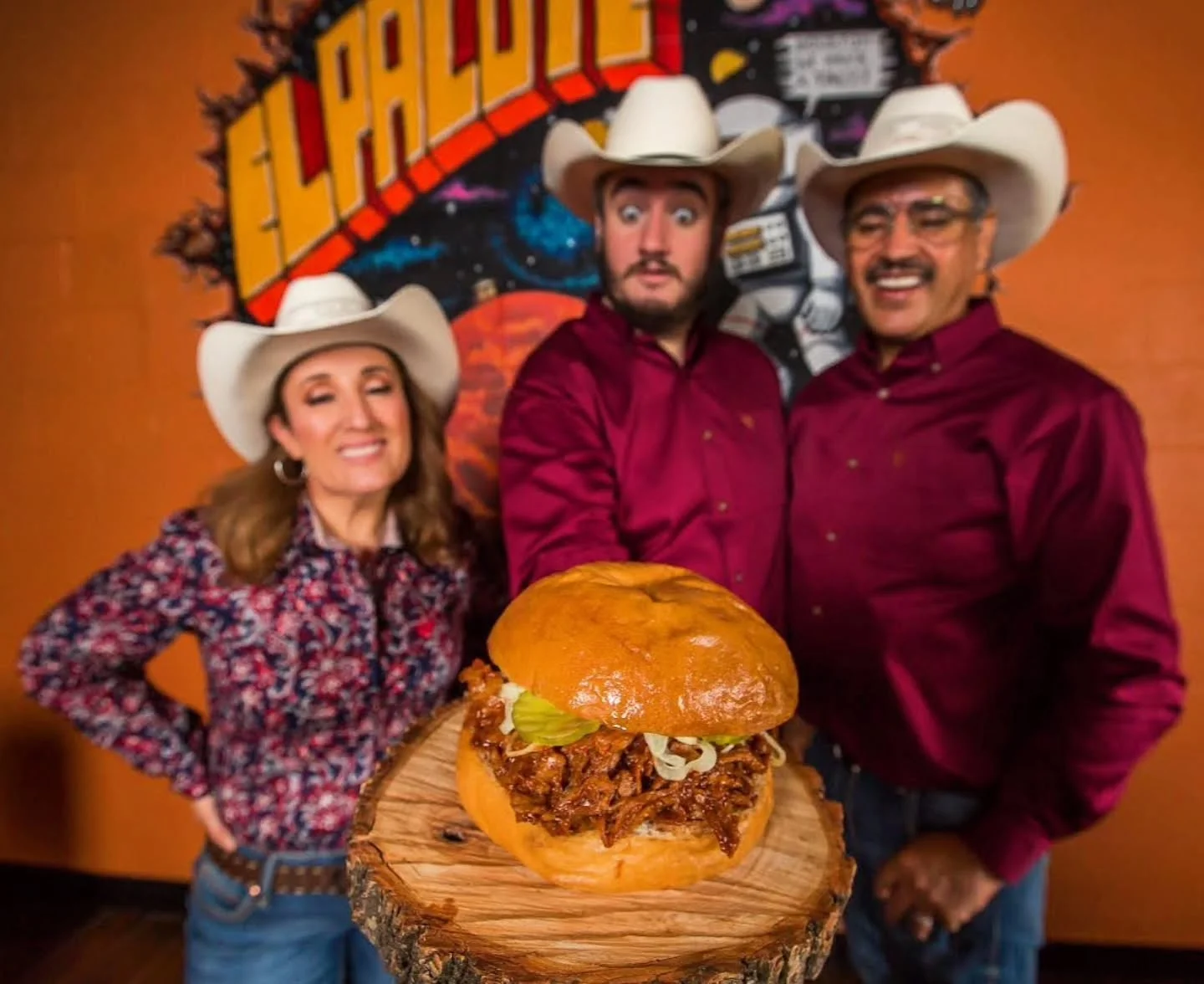 Three people presenting a delicious pulled pork sandwich in a vibrant restaurant setting.