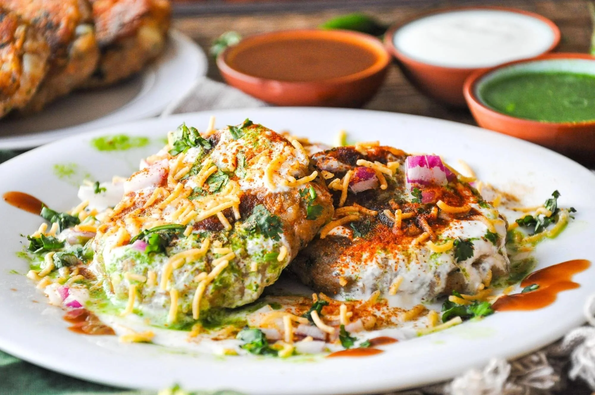 Two servings of a savory Indian street food dish, topped with sauces, yogurt, chopped onions, and garnished with cilantro and crispy elements.