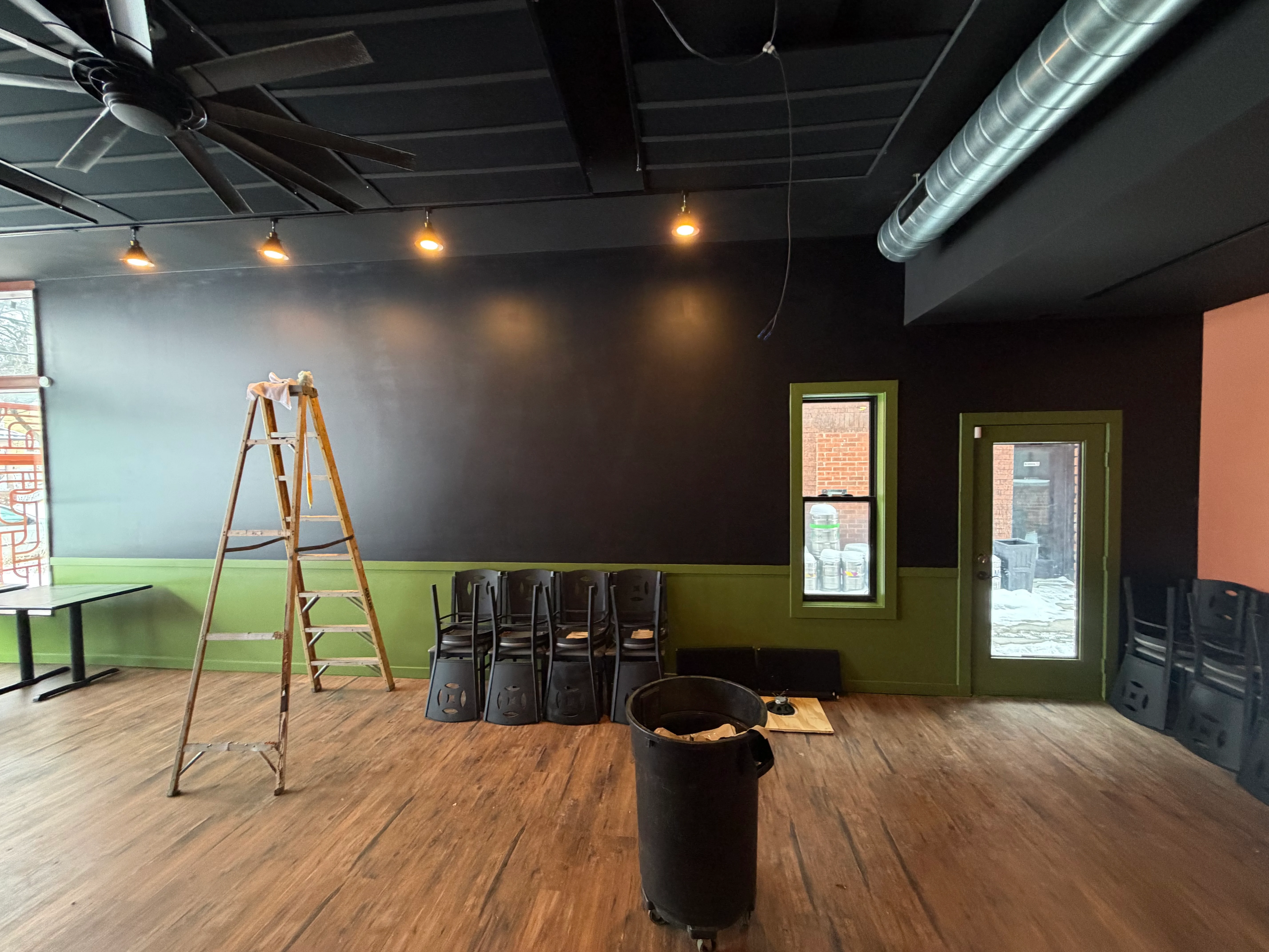 An unfinished dining space featuring a ladder and stacked chairs. The walls are painted in two tones, and there's a window providing natural light.