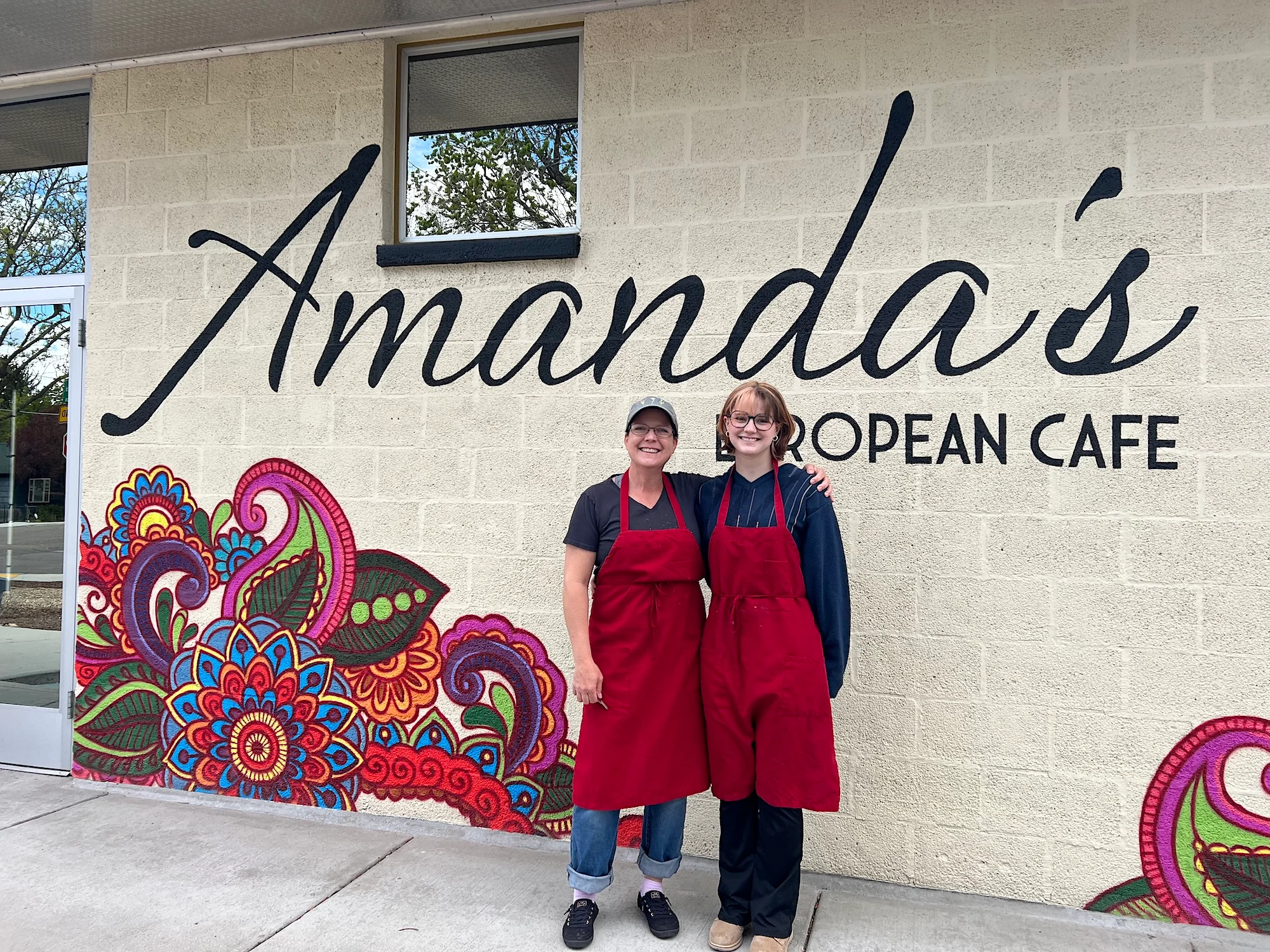 Two individuals wearing red aprons stand in front of a colorful mural depicting intricate patterns, alongside a sign for a European cafe.
