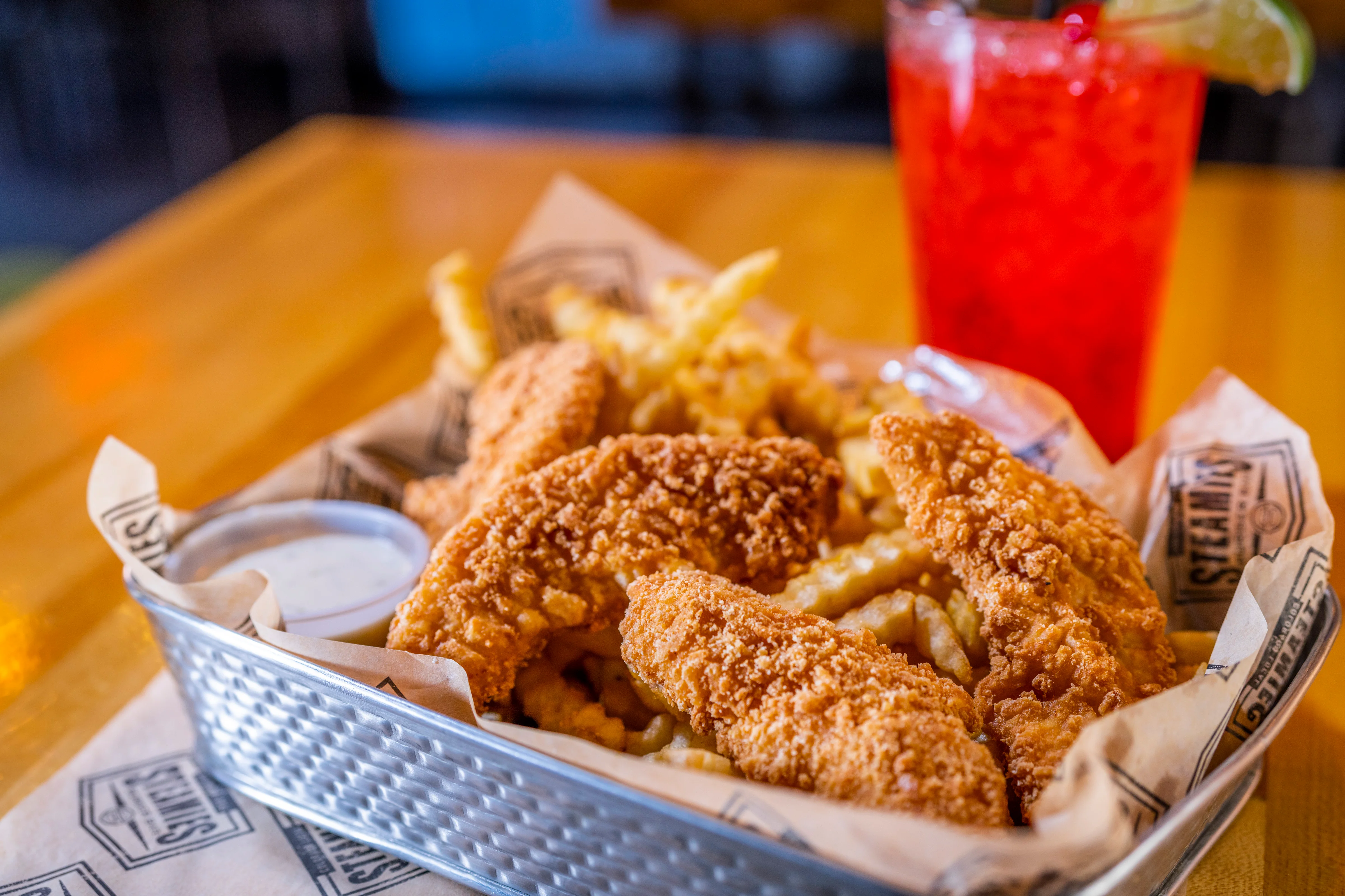 A basket of crispy fried chicken tenders served with crinkle-cut fries and a side of dipping sauce. A refreshing drink is in the background.