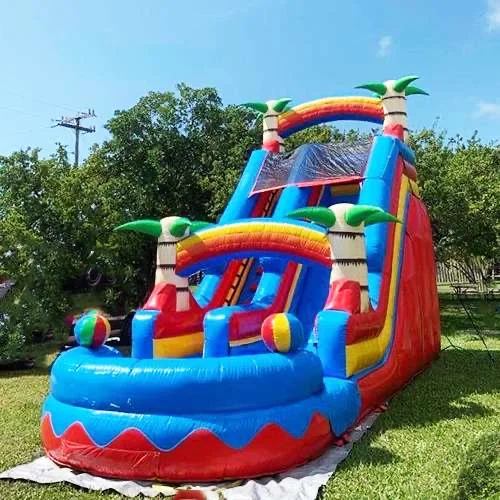 A colorful inflatable water slide with palm trees and beach ball decorations, set on a grassy area.