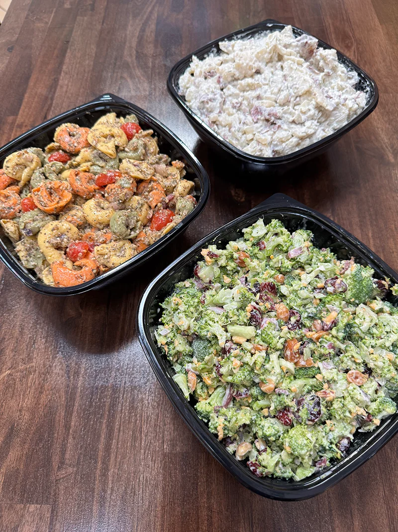 A table displays three black containers: creamy red potato salad, almond pesto tortellini, and a colorful broccoli salad.