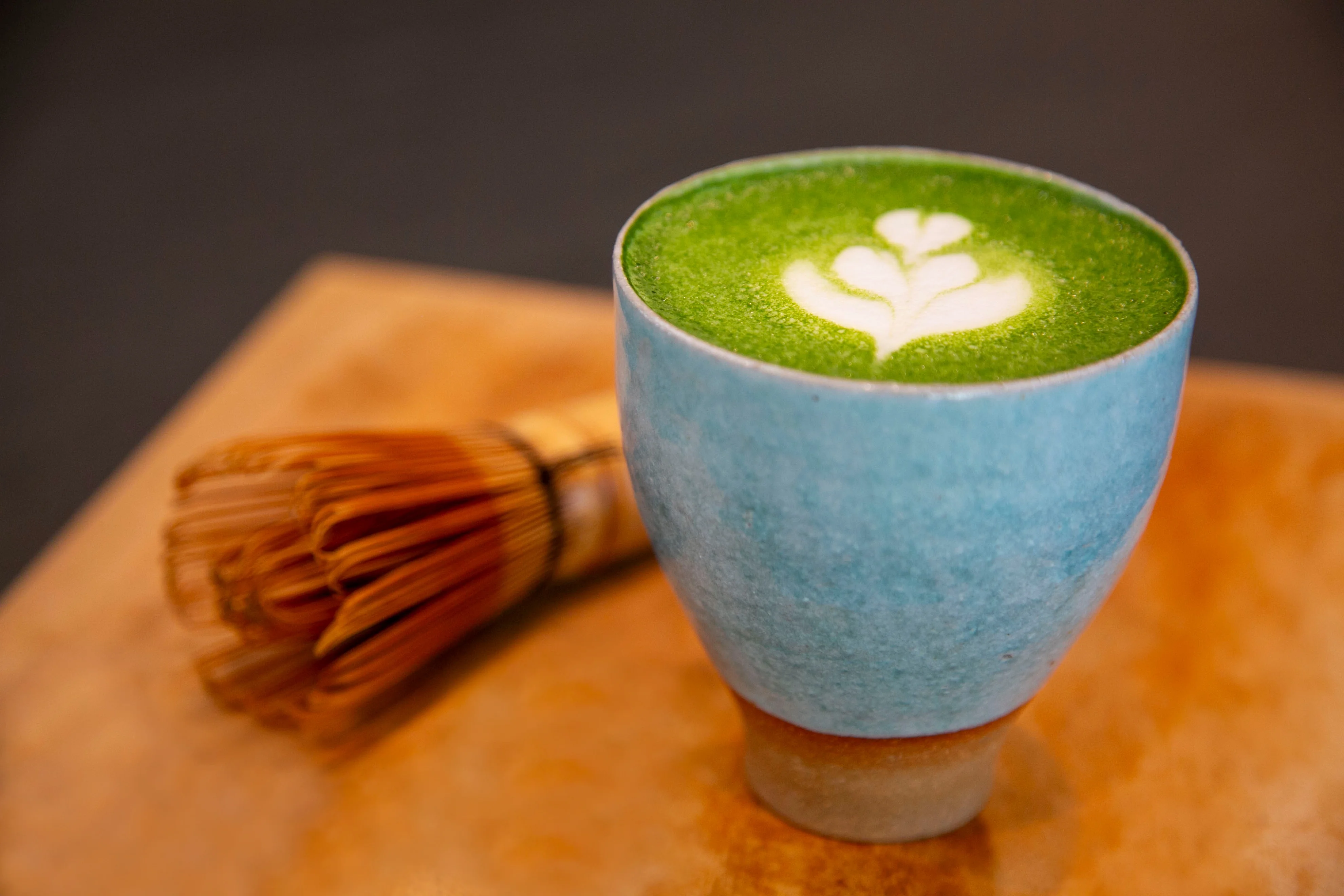 A vibrant green latte topped with a delicate leaf design, served in a blue ceramic cup alongside a traditional bamboo whisk.
