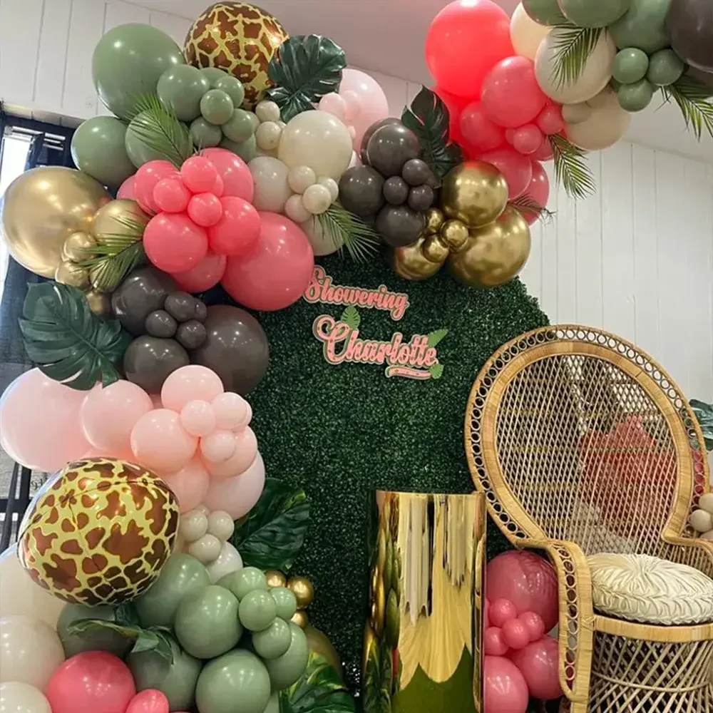 A festive balloon decoration featuring various colors and textures, including greens, pinks, and gold, arranged around a backdrop with the text 'Showering Charlotte.' A rattan chair and a reflective golden column are also present.