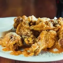 A plate of crispy chicken wings coated in a spicy sauce and sprinkled with cheese, served with a small cup of dipping sauce.