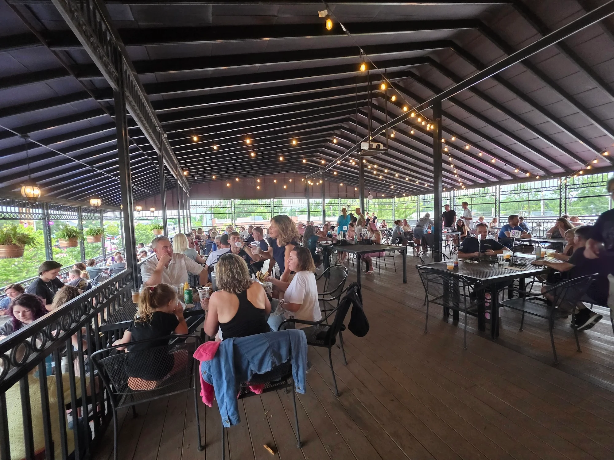 A lively outdoor dining area filled with guests enjoying their meals under string lights, in a spacious covered setting.