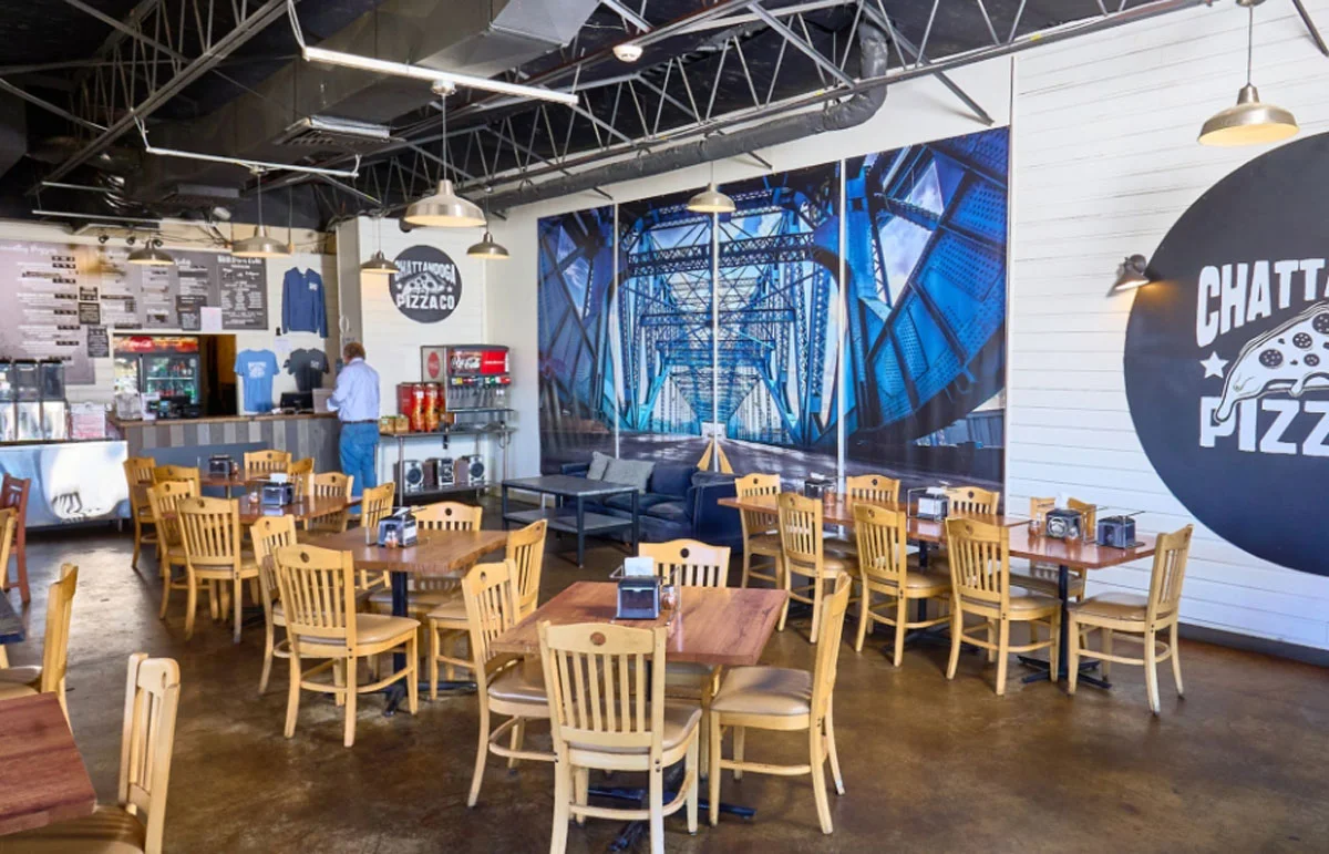 An interior view of a pizza restaurant featuring wooden tables and chairs, along with a decorative mural and a service counter.