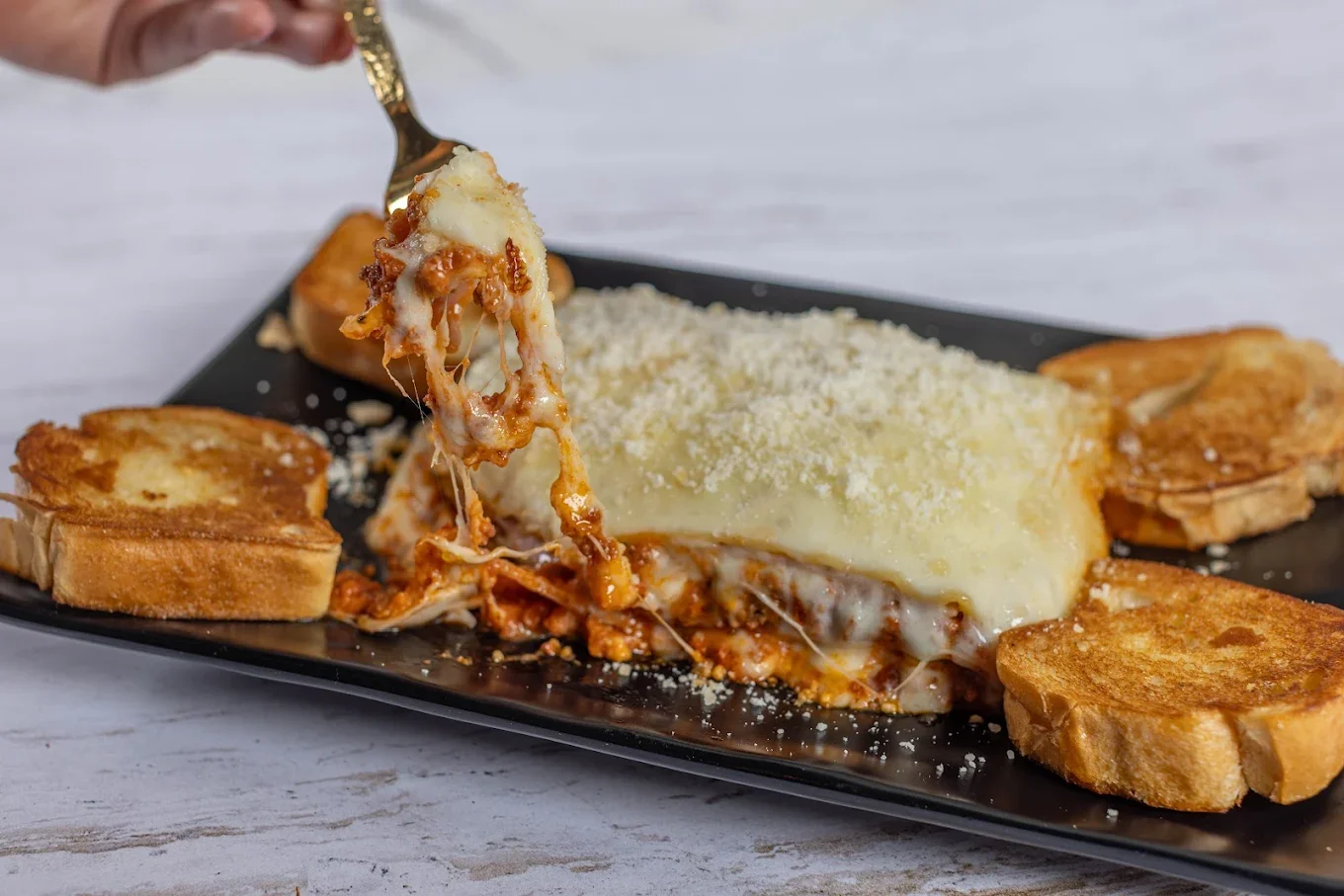A close-up of a hearty portion of baked lasagna with meat sauce, served with garlic bread, with a fork pulling away a piece to create a delicious cheese pull.