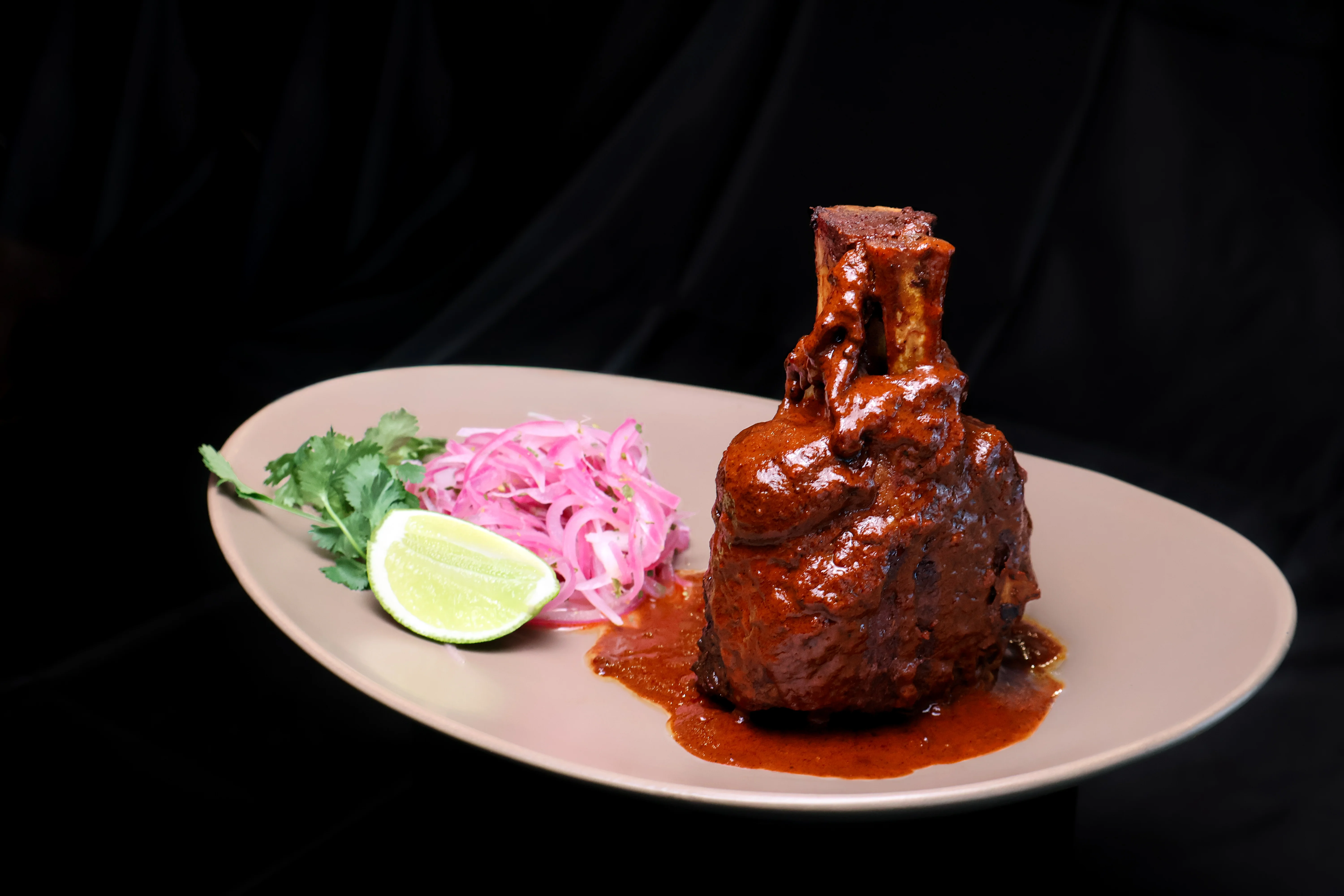 A beautifully presented dish featuring a large piece of meat covered in rich sauce, accompanied by pickled onions, cilantro, and a lime slice on a beige plate.